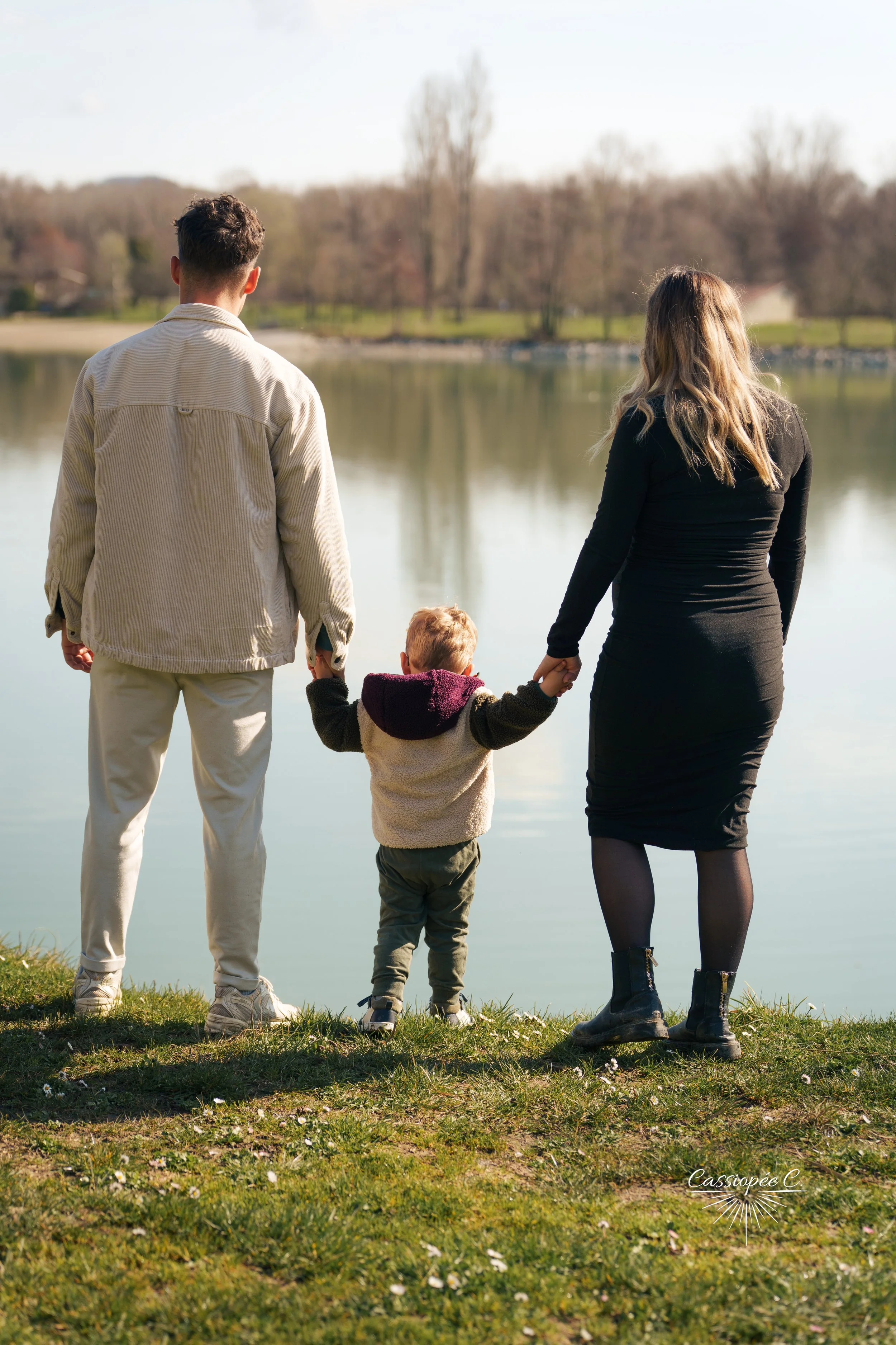 Une famille de trois personnes, un homme, une femme, et un enfant, se tient au bord d'un lac, main dans la main, avec un paysage de arbres et d'eau en arrière-plan.