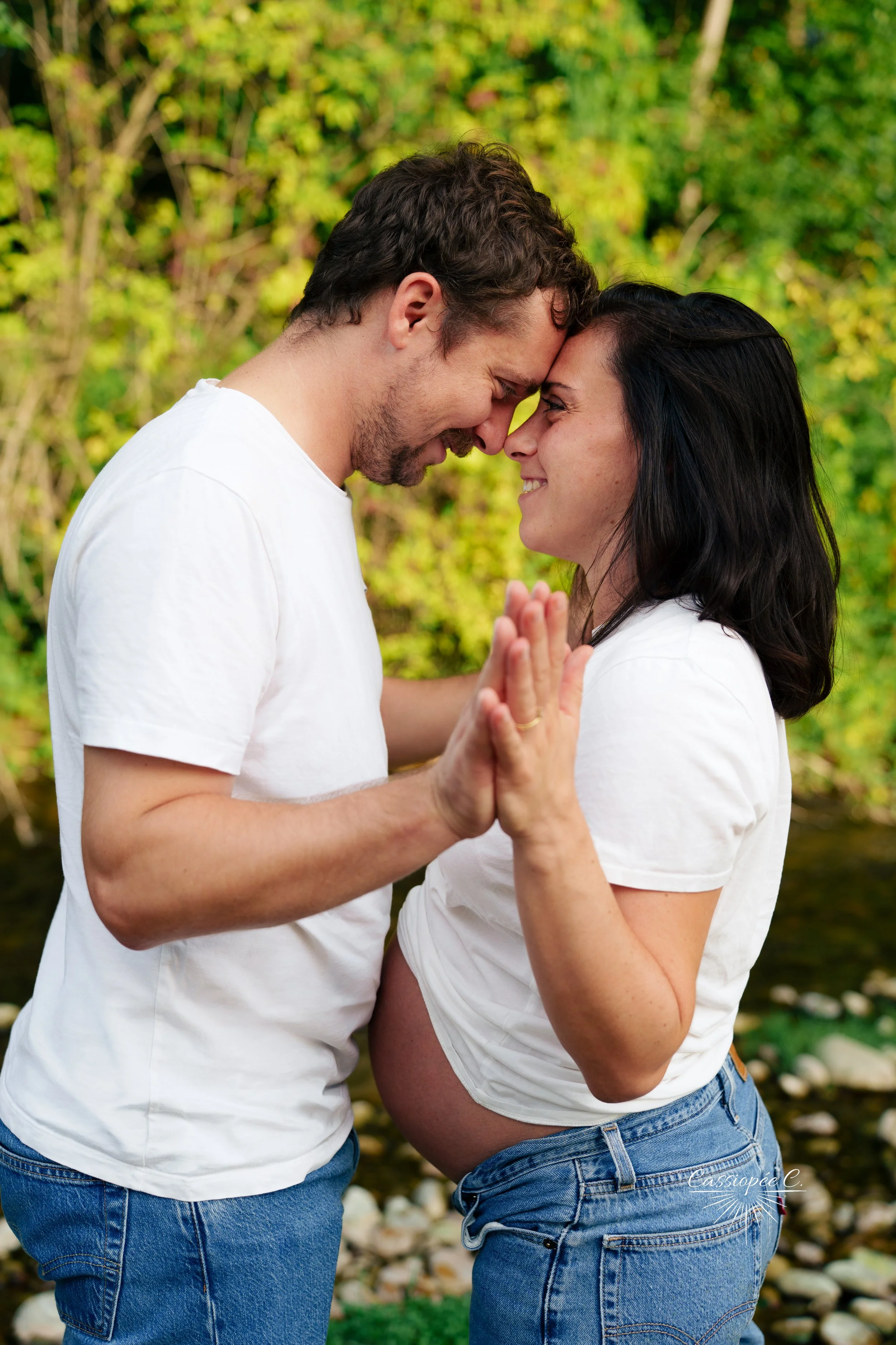 Un couple s'appuyant le front l'un contre l'autre, mains jointes à hauteur de leur ventre, en plein air avec un fond de feuillage vert.