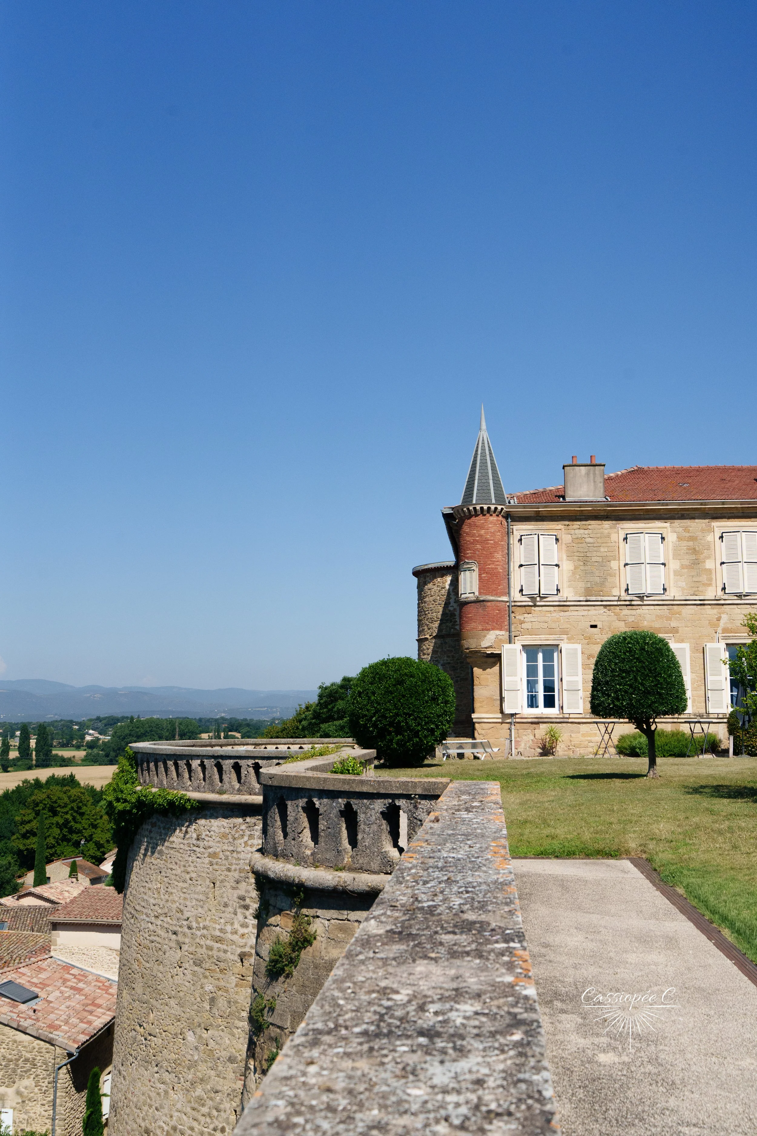 Vue d'un vieux château avec un mur en pierre, une tour pointue, des fenêtres à volets blancs et un jardin bien entretenu avec deux arbres arrondis, sous un ciel clair et bleu.