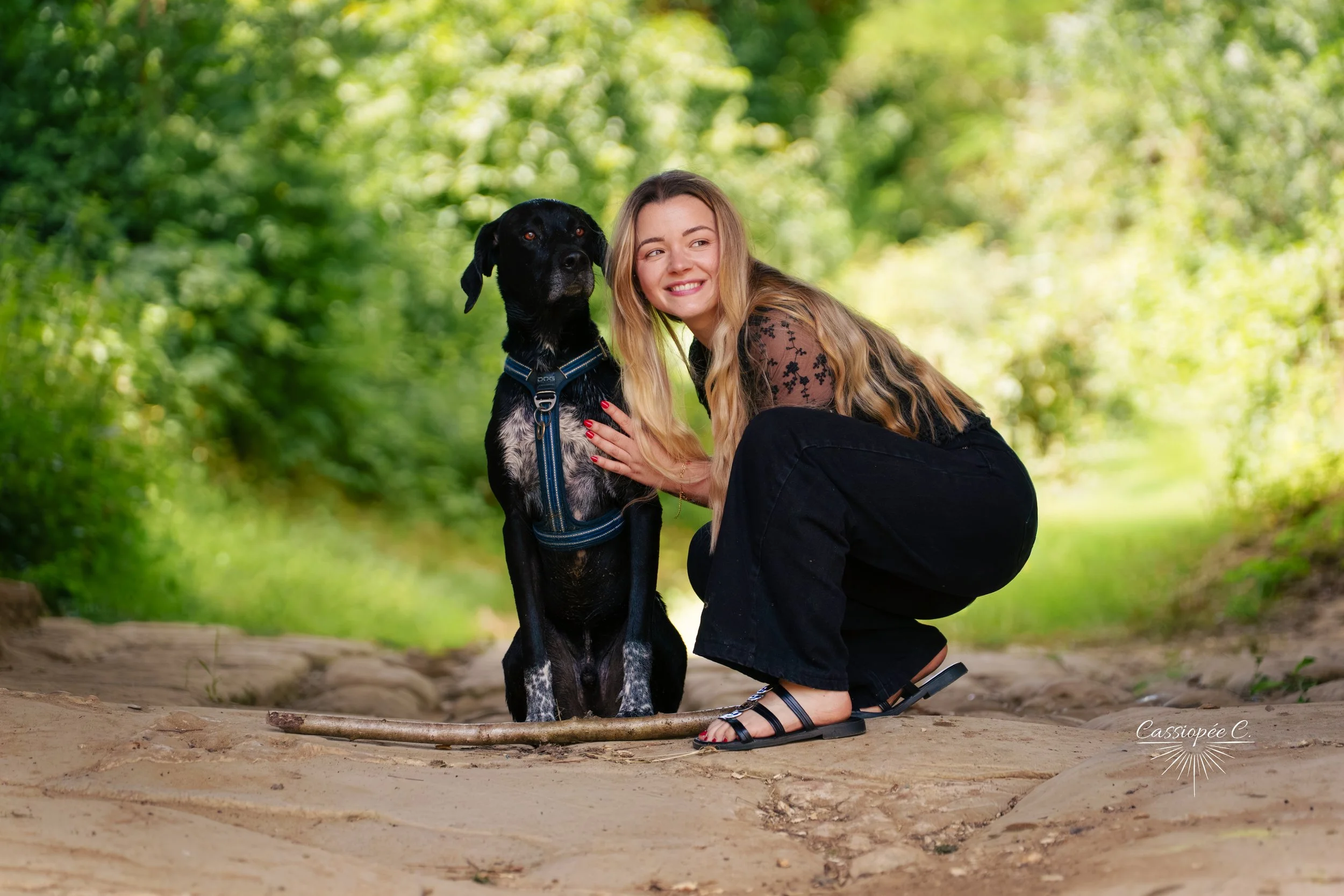 Une femme souriante avec de longs cheveux blonds, portant une robe noire, est agenouillée à côté d'un chien noir à pattes tachetées, dans un environnement forestier vertueux, au sol en terre et bois.