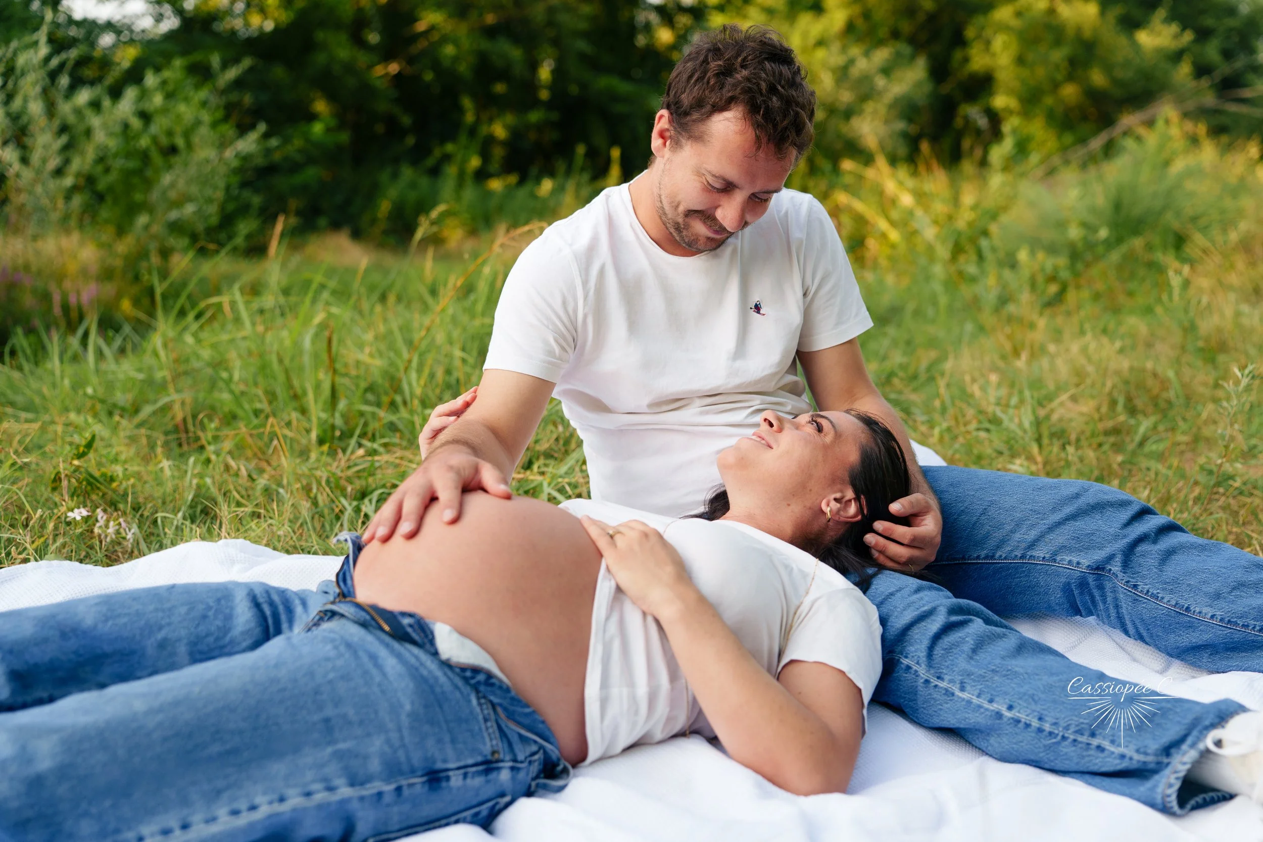 Un homme caresse le ventre de sa femme allongée sur une couverture dans un environnement naturel avec des arbres et de l'herbe verte.