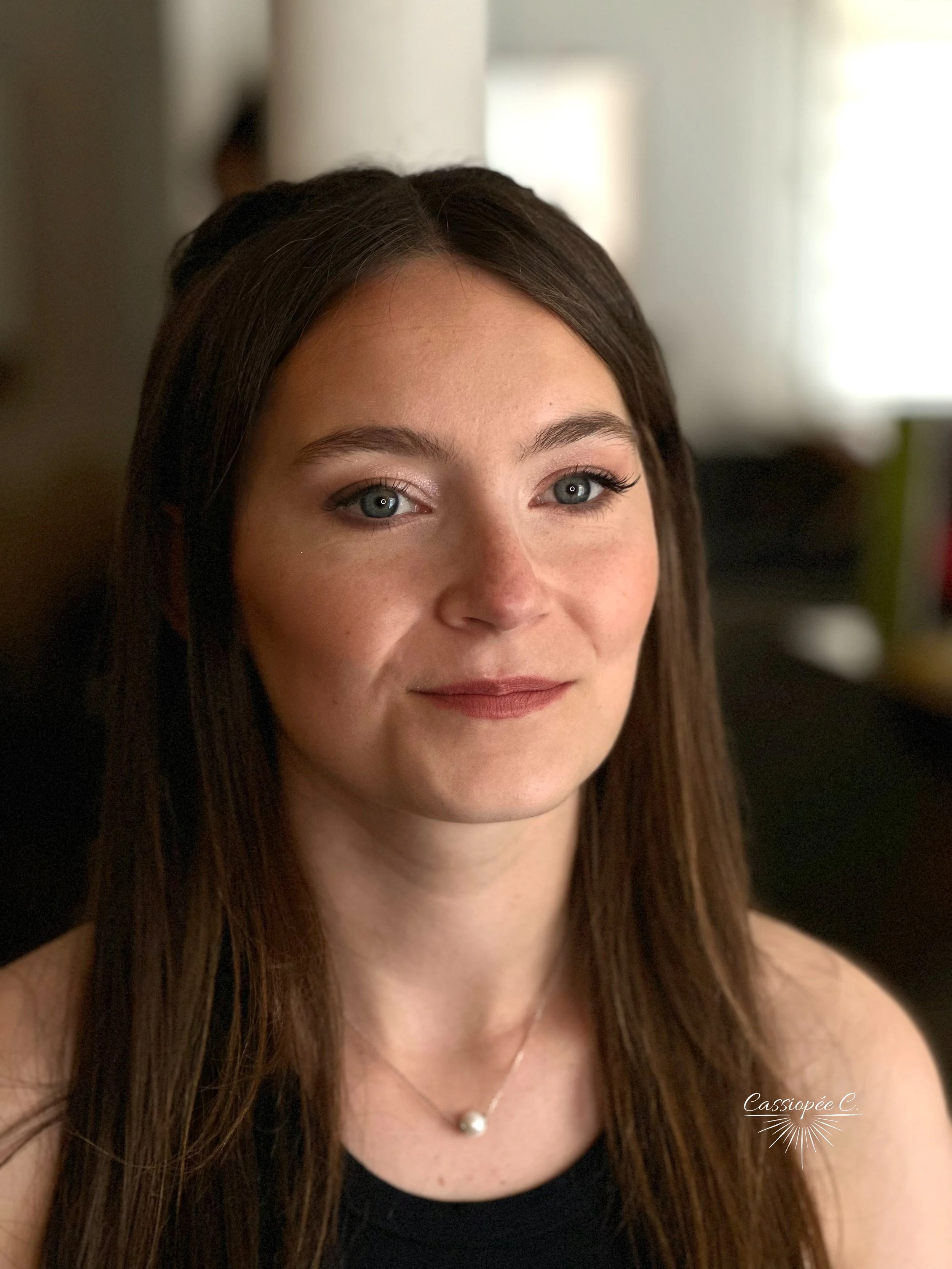 Jeune femme avec les cheveux longs, yeux bleus, souriant légèrement, dans un intérieur lumineux avec un maquillage pour un mariage.