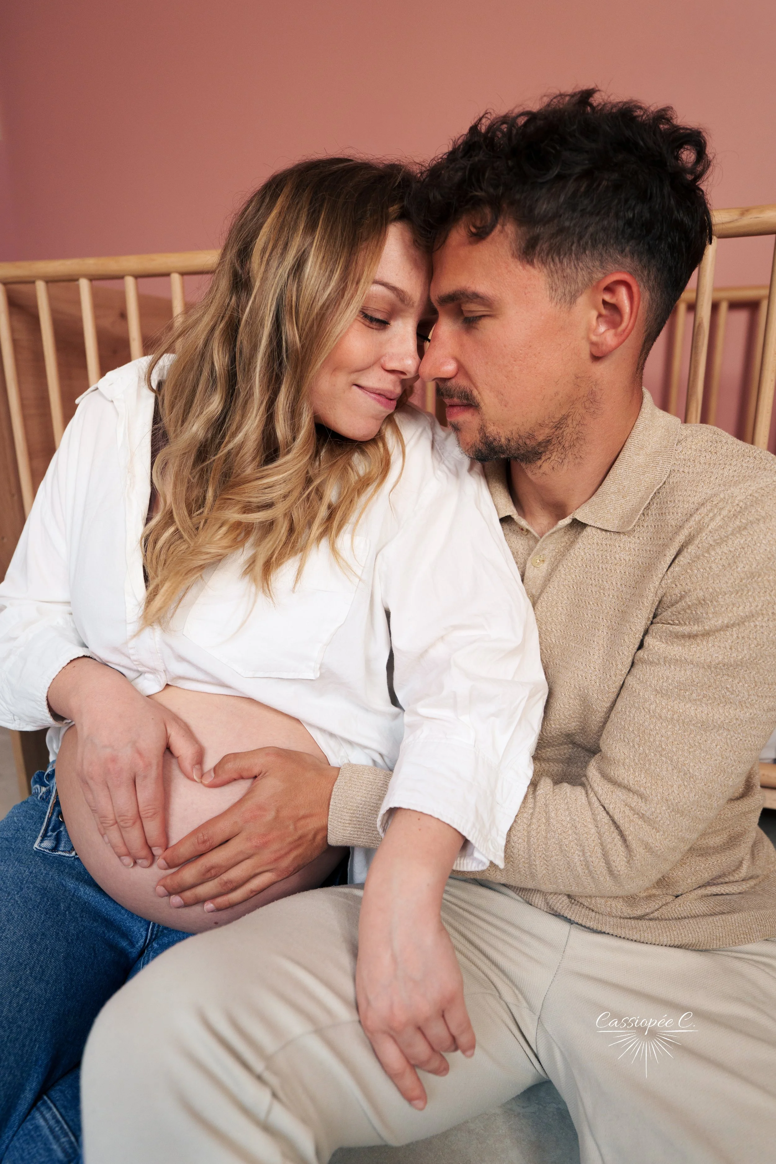 Un couple se tient proche, la femme enceinte tenant son ventre tandis que l'homme le soutient, dans une pièce avec un fond rose et un lit en bois.