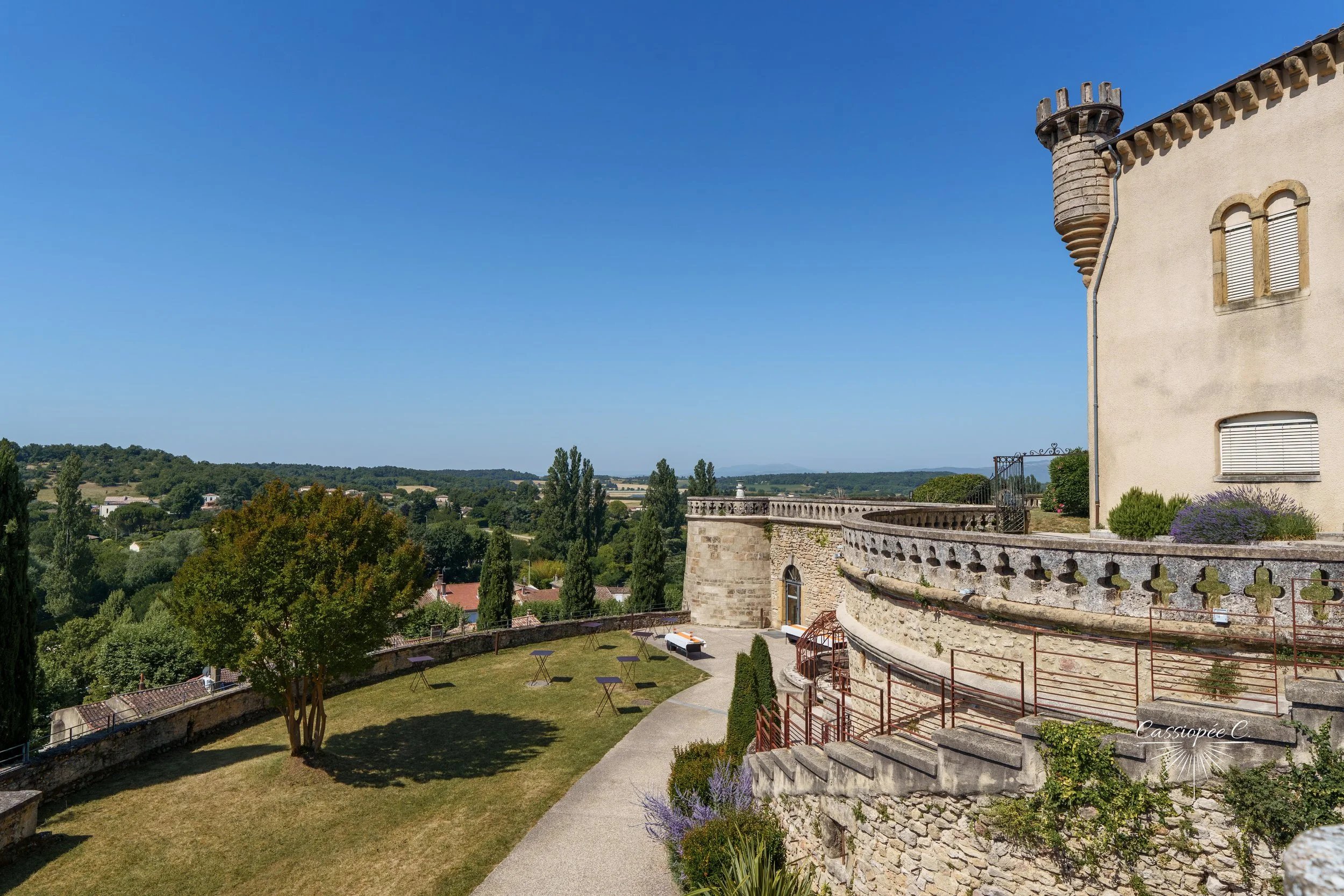 Une cour extérieure d'un château en pierre avec terrasse, arbres, et un paysage rural avec des collines en arrière-plan, sous un ciel bleu clair.