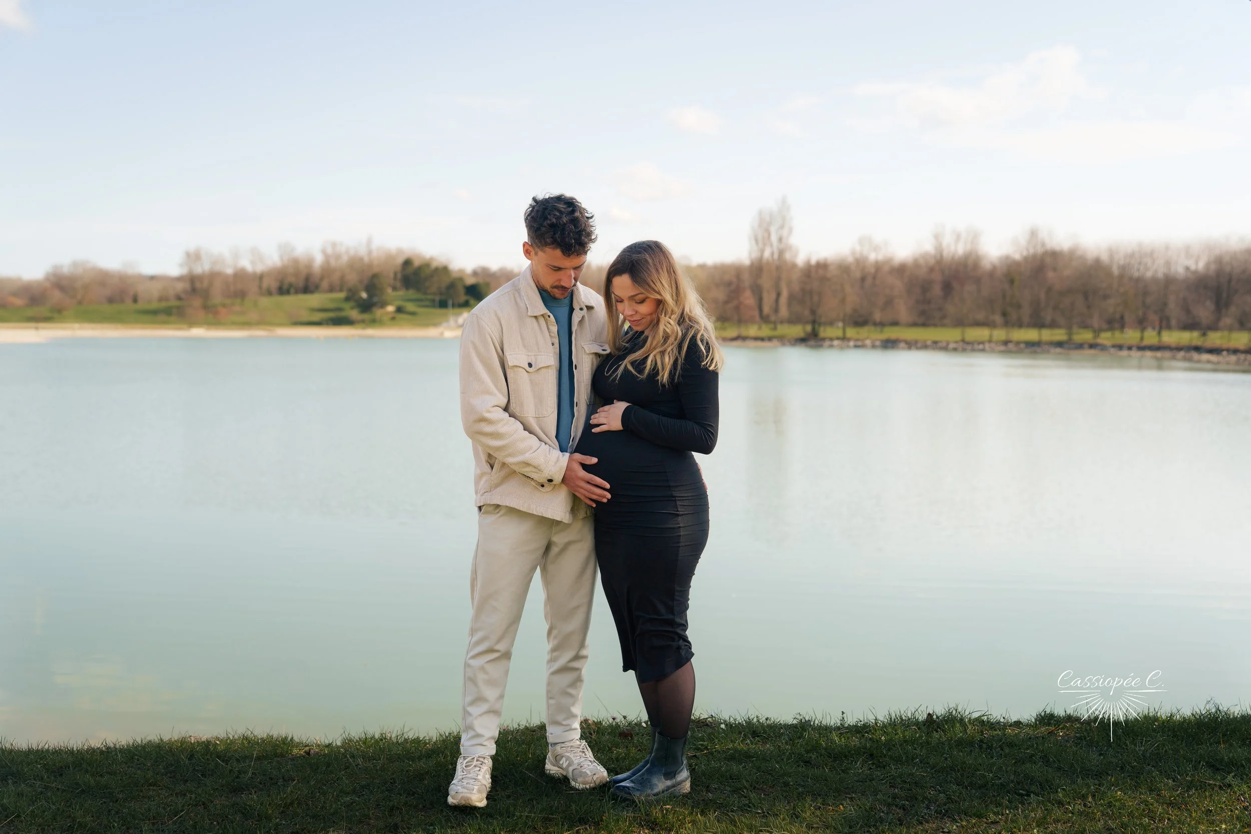 Un couple enceinte se tient près d'un lac par une journée ensoleillée, tous deux regardant leur ventre. Paysage naturel avec arbres et ciel bleu en arrière-plan.