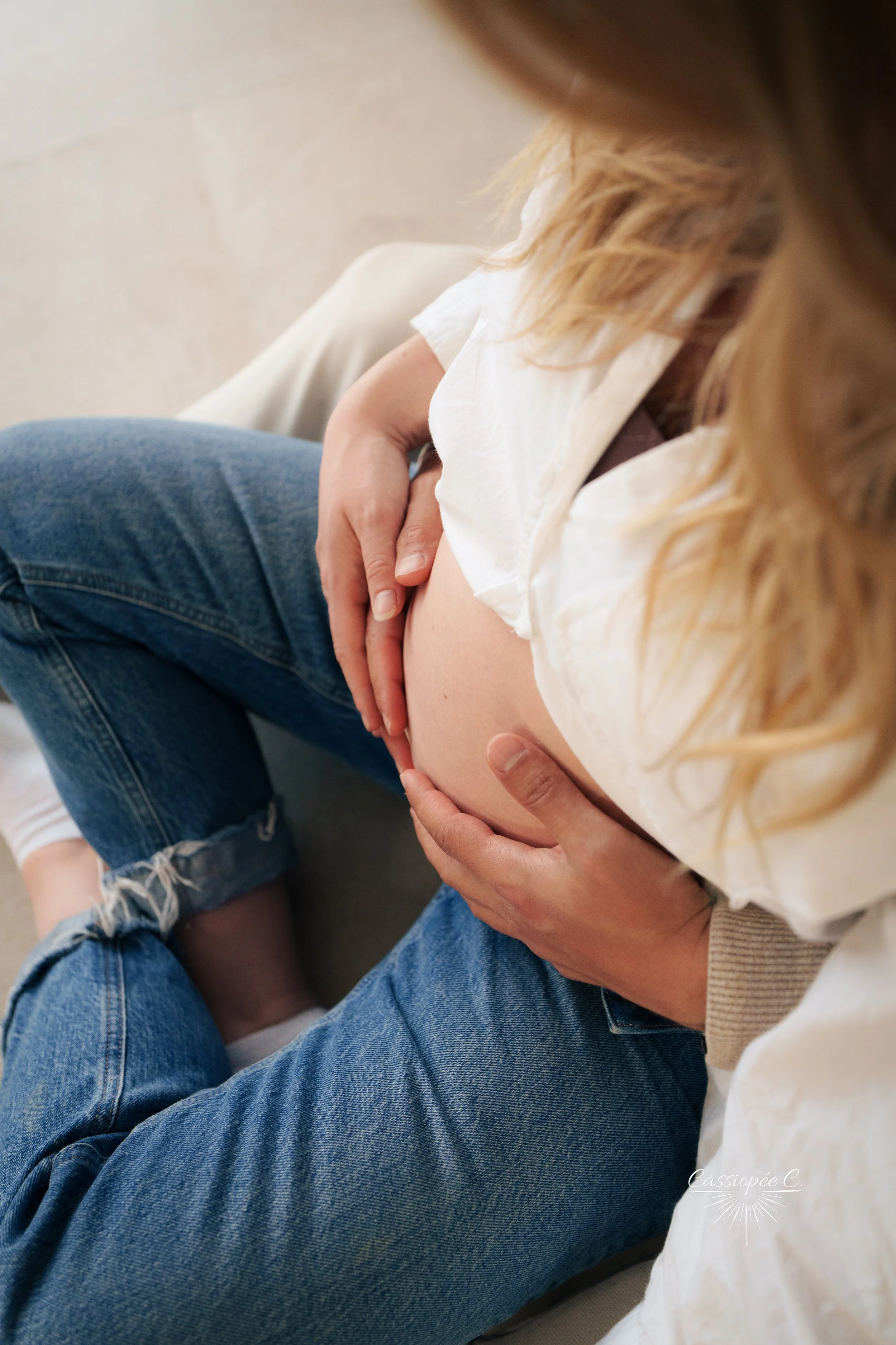 Une femme enceinte assise, tenant son ventre avec ses mains, portant un t-shirt blanc et un jean bleu déchiré.