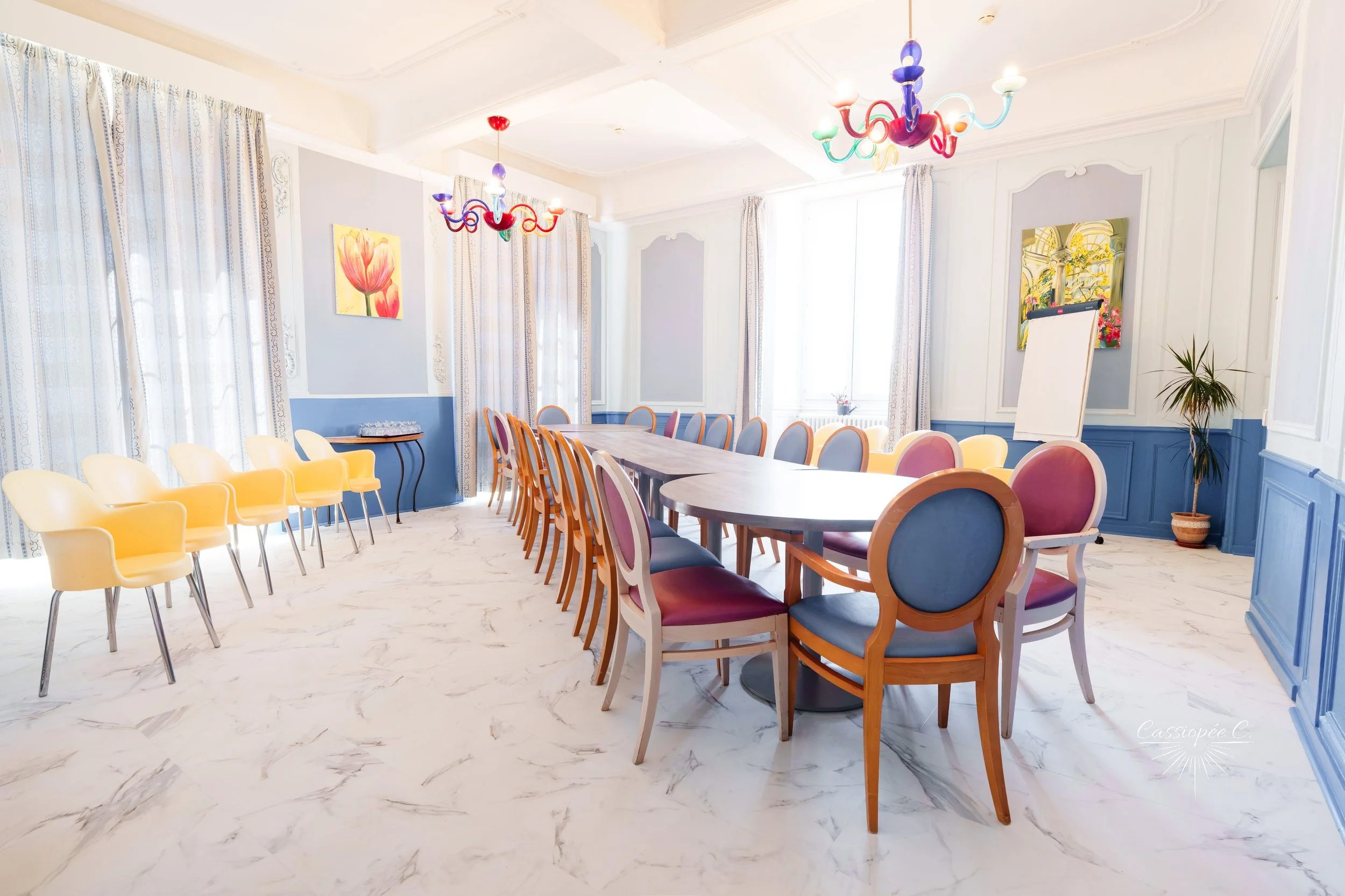 Salle de réunion bien éclairée avec plusieurs chaises colorées autour d'une grande table en bois, murs ornés de tableaux floraux, deux lustres colorés suspendus au plafond, et une grande fenêtre avec des rideaux, décoration élégante et lumineuse.