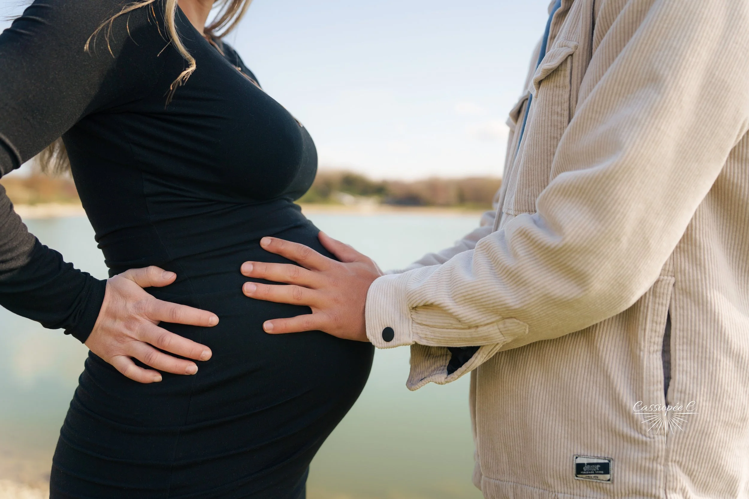 Une femme enceinte habillée en noir et un homme habillé en beige se tiennent face à face près d'une rivière ou d'un lac, avec leurs mains posées sur le ventre de la femme. Le contexte est naturel avec un ciel clair en arrière-plan.