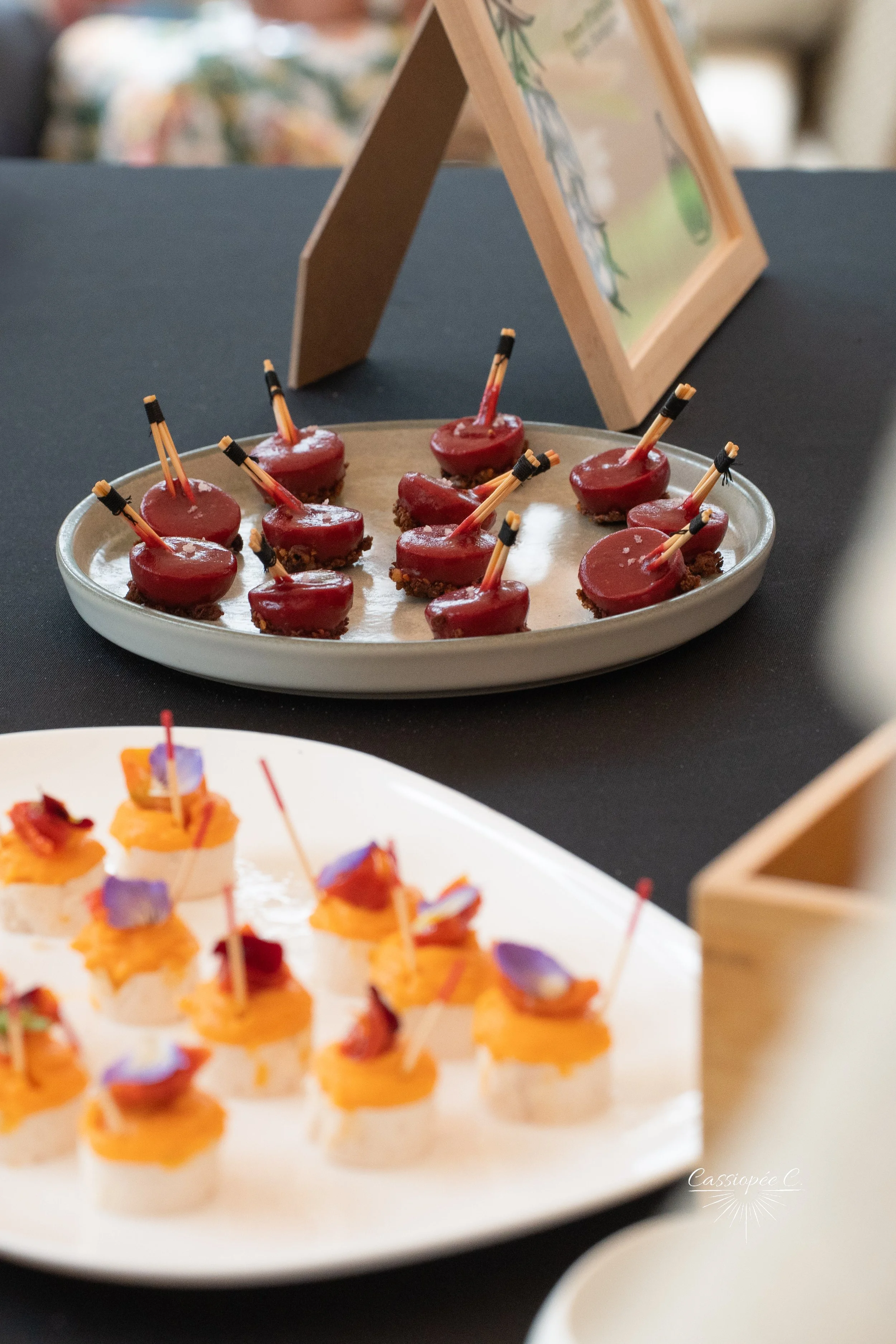 Petits fours sucrés présentés sur deux plateaux, décorés avec des cure-dents et des fleurs comestibles, avec un cadre en bois en arrière-plan.