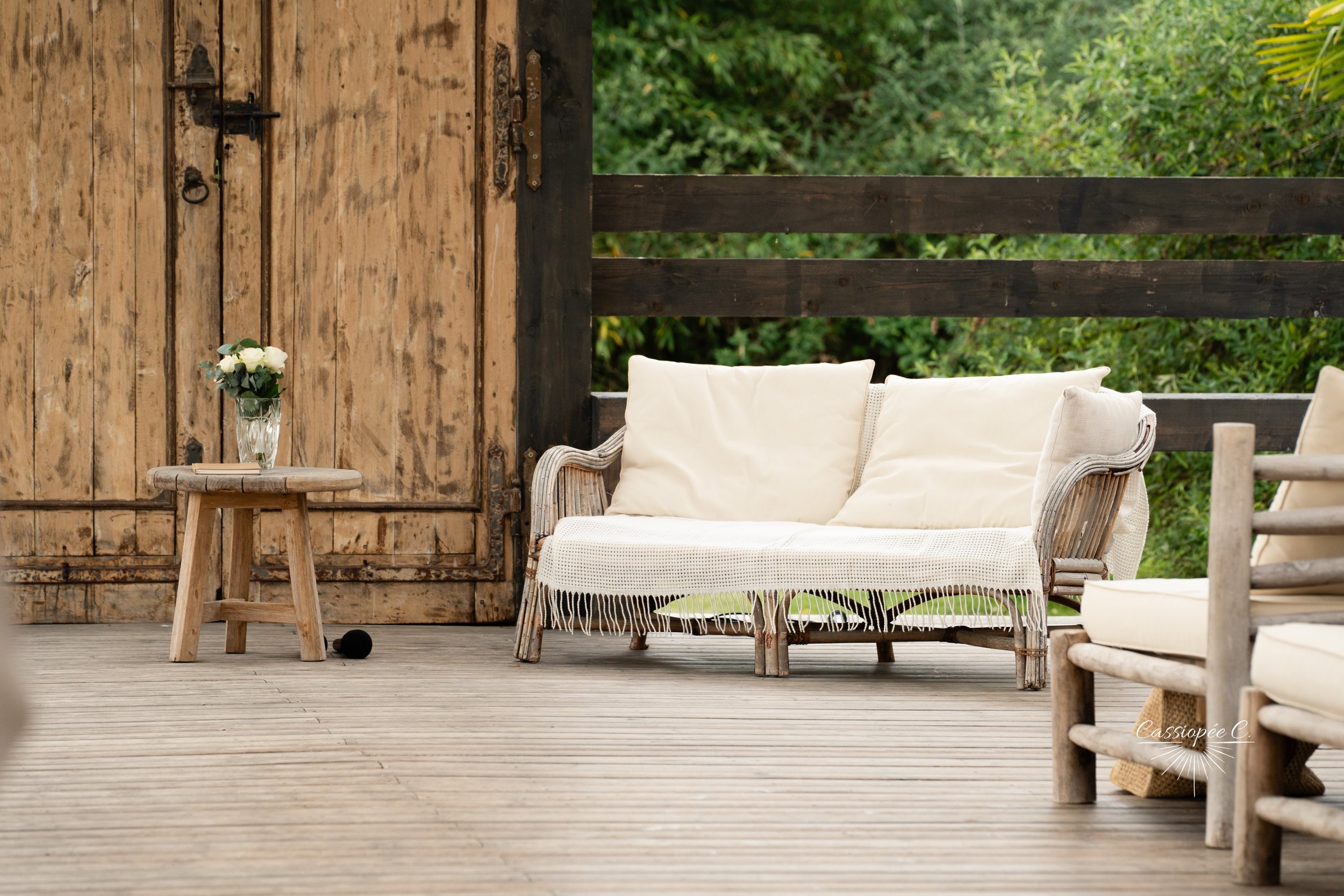Une terrasse en bois avec un canapé en rotin blanc avec des coussins, un petit tabouret avec un vase de fleurs, et un fauteuil en bois avec des coussins, entourée de verdure.