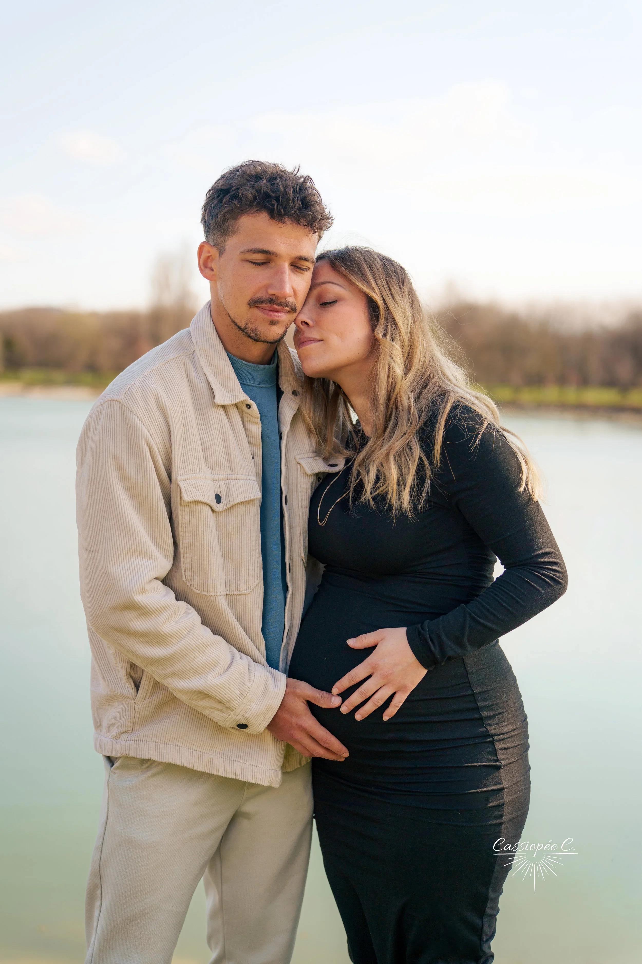 Un couple, un homme et une femme, se tiennent près d'une rivière, avec des paysages naturels en arrière-plan. La femme enceinte a la main sur son ventre, et ils ont une expression d'affection et de calme.