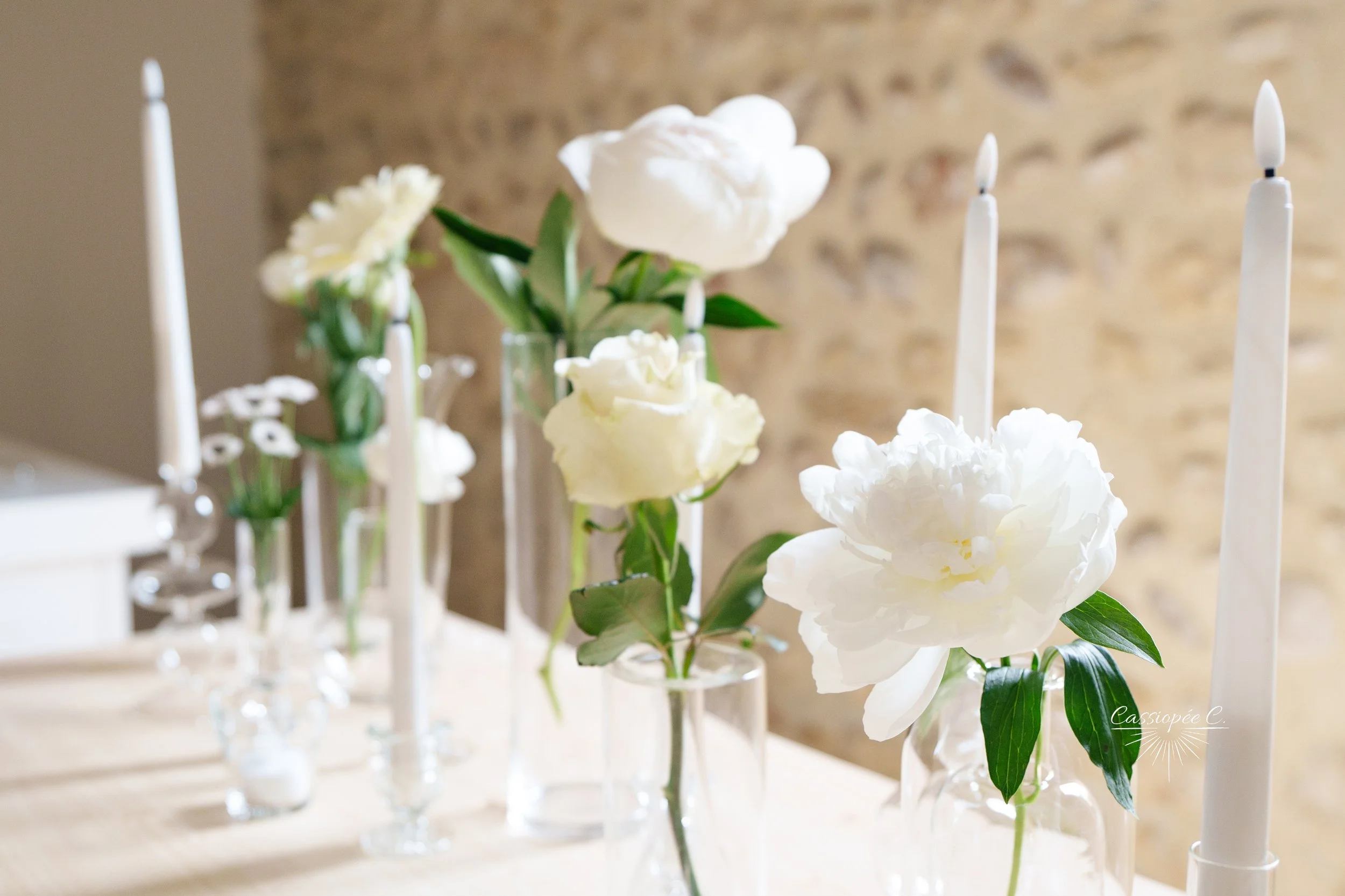 Vase avec des fleurs blanches, notamment des pivoines, accompagnées de bougies allumées dans des chandeliers blancs, disposés sur une table, avec un mur en pierre en arrière-plan.
