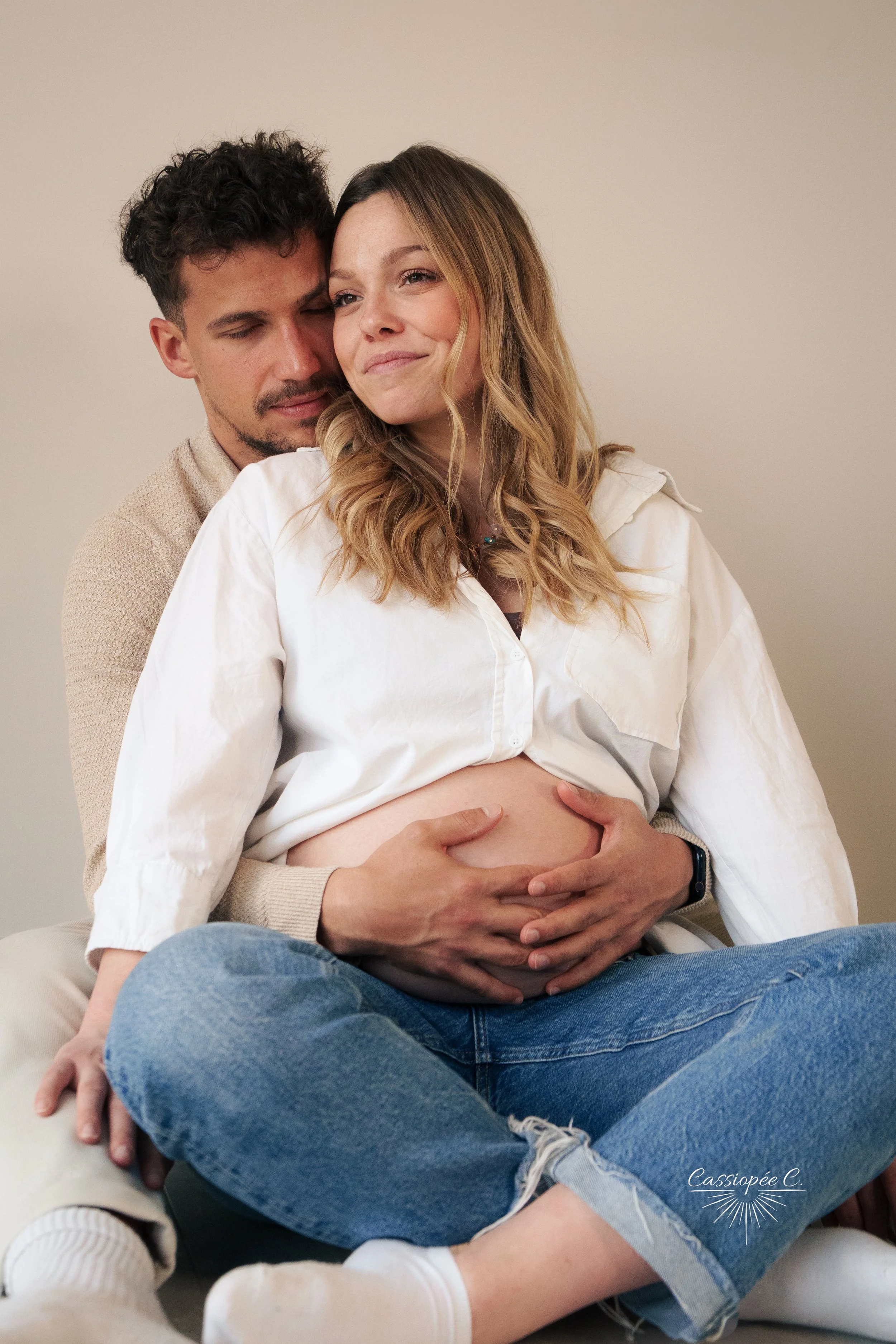 Un couple assis, la femme enceinte, tenant son ventre, avec l'homme derrière elle, tous deux souriants et regardant dans des directions différentes.
