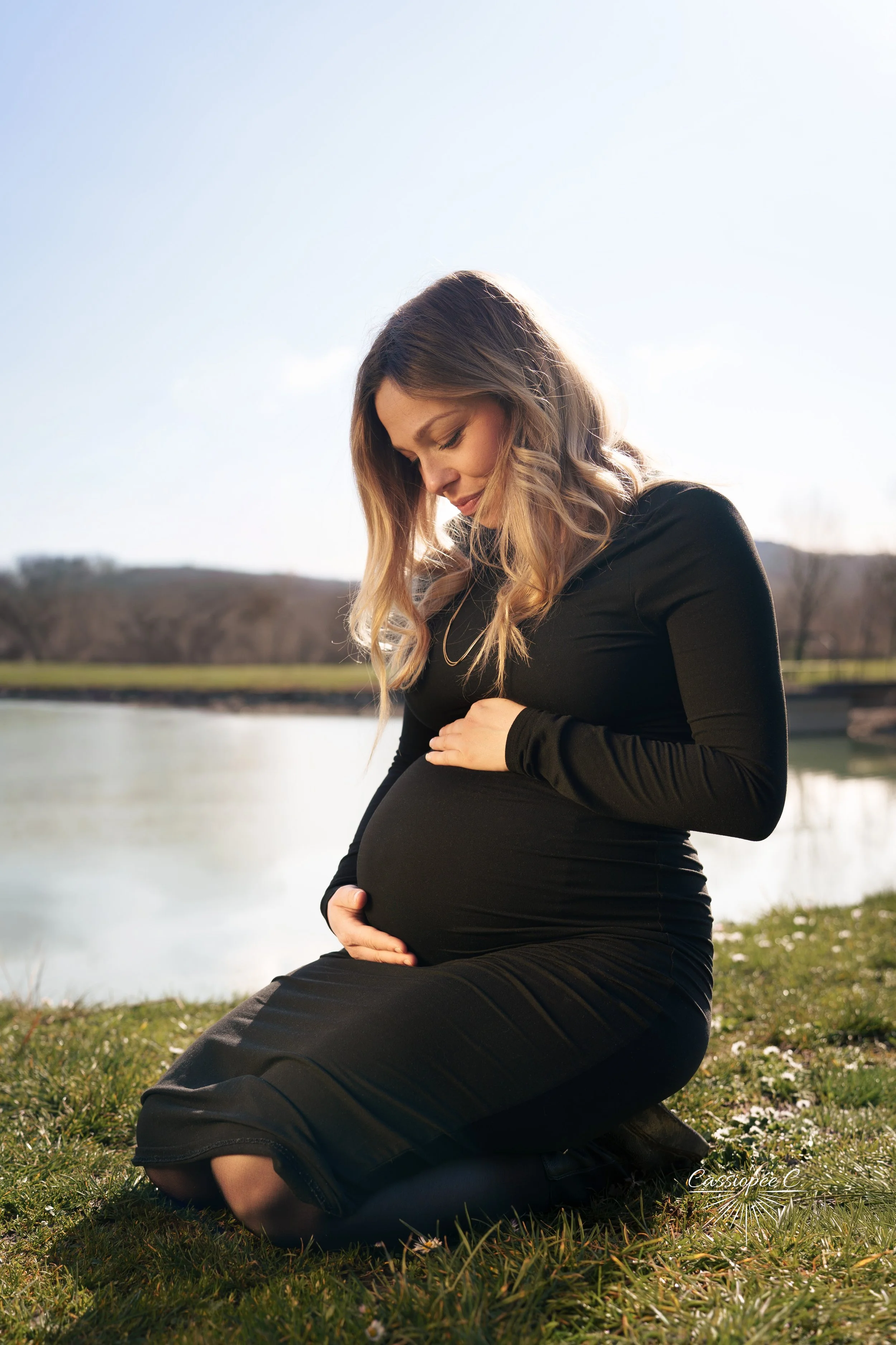Une femme enceinte assise sur l'herbe près d'un lac, tenant son ventre avec ses mains et regardant vers le bas, dans un environnement naturel en plein air. Il fait jour avec un ciel clair.