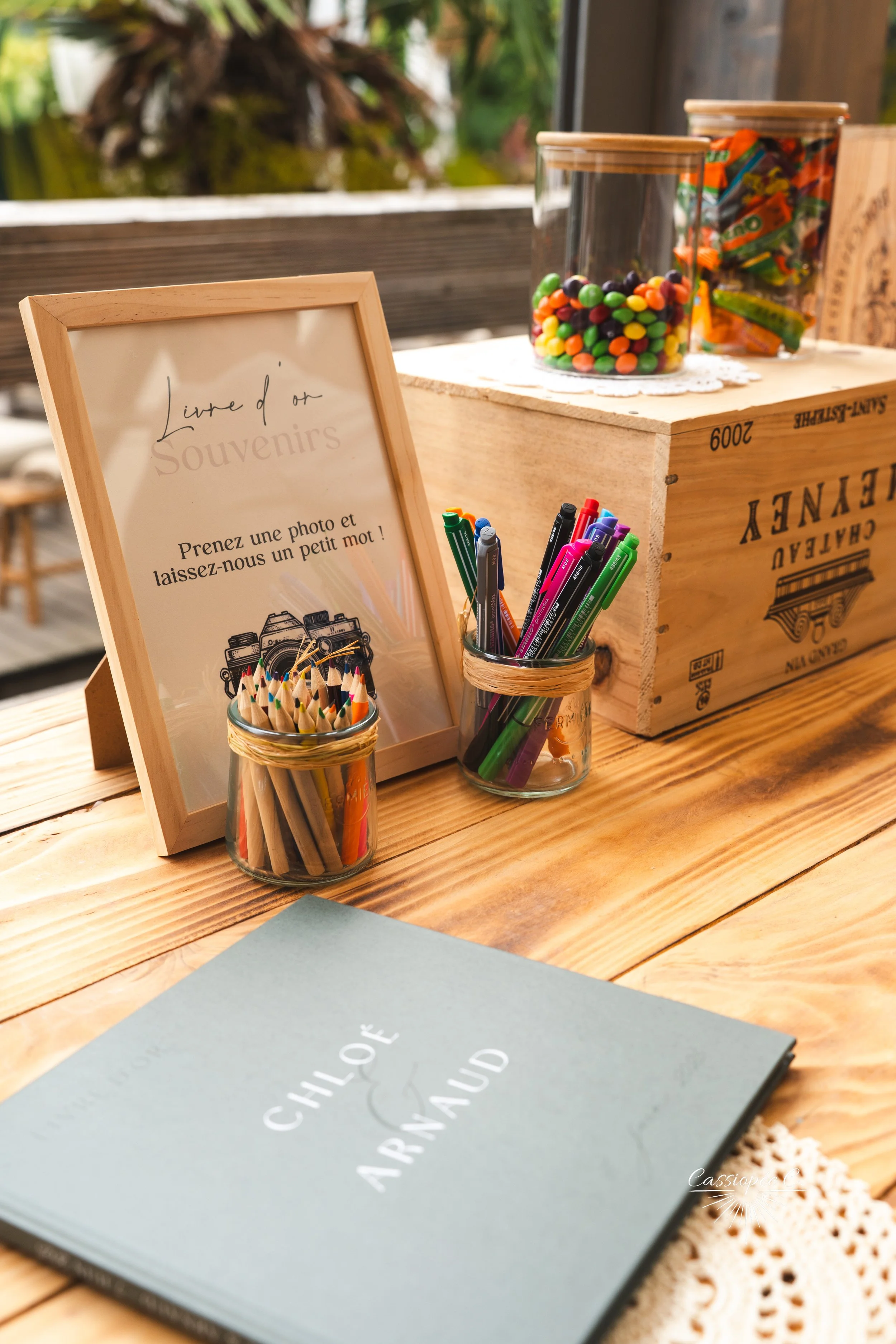 Table en bois avec un cadre photo, deux petits pots remplis de stylos, un cahier vert clair intitulé "Chloé Arnaud" et deux bocaux en verre contenant des bonbons et des stylos, dans un espace en plein air.