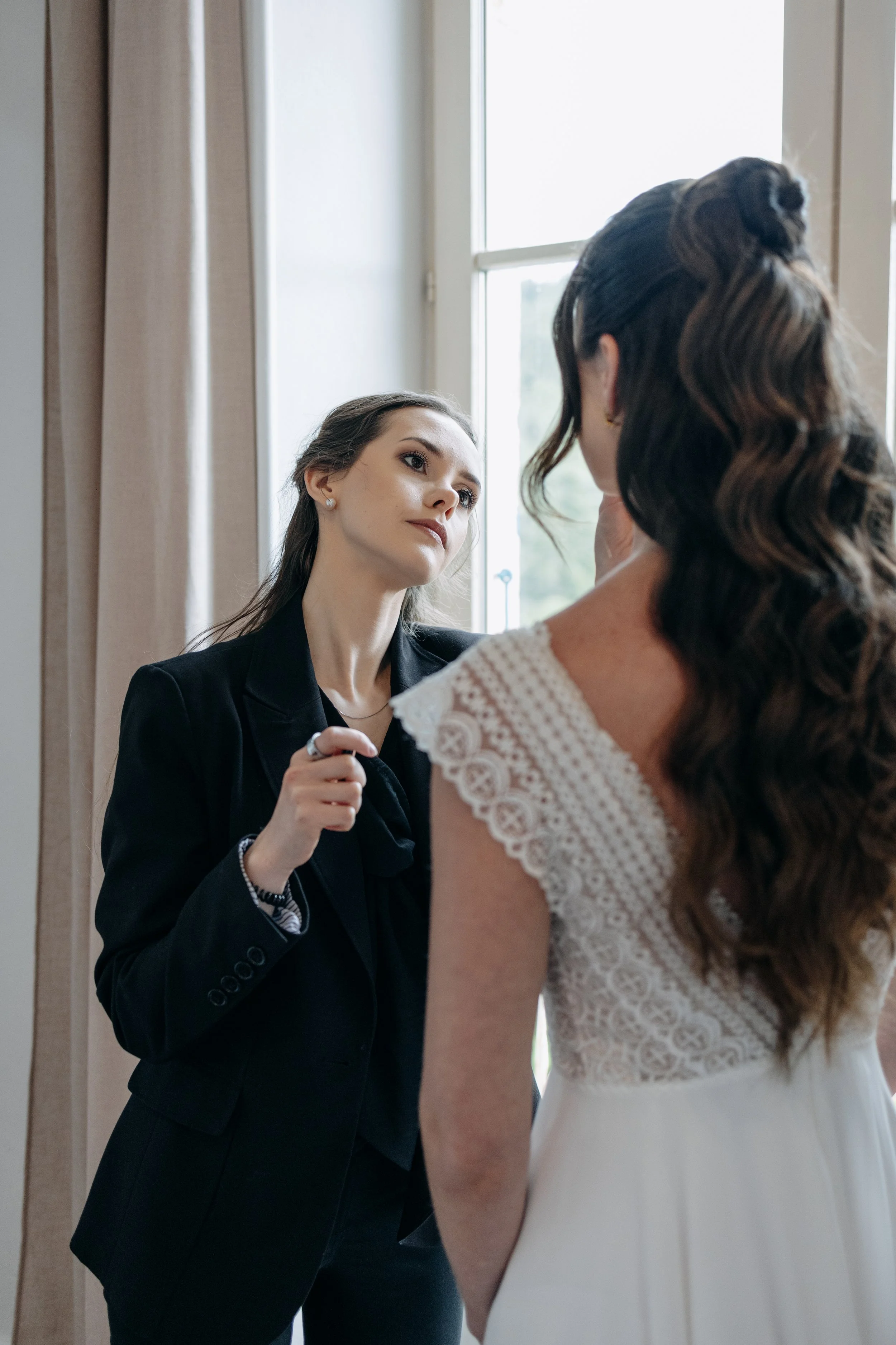 Une maquilleuse en vêtements noirs sublime une mariée lors de la préparation avant le mariage. La mariée porte une robe blanche avec dentelle et a de longs cheveux bruns bouclés.