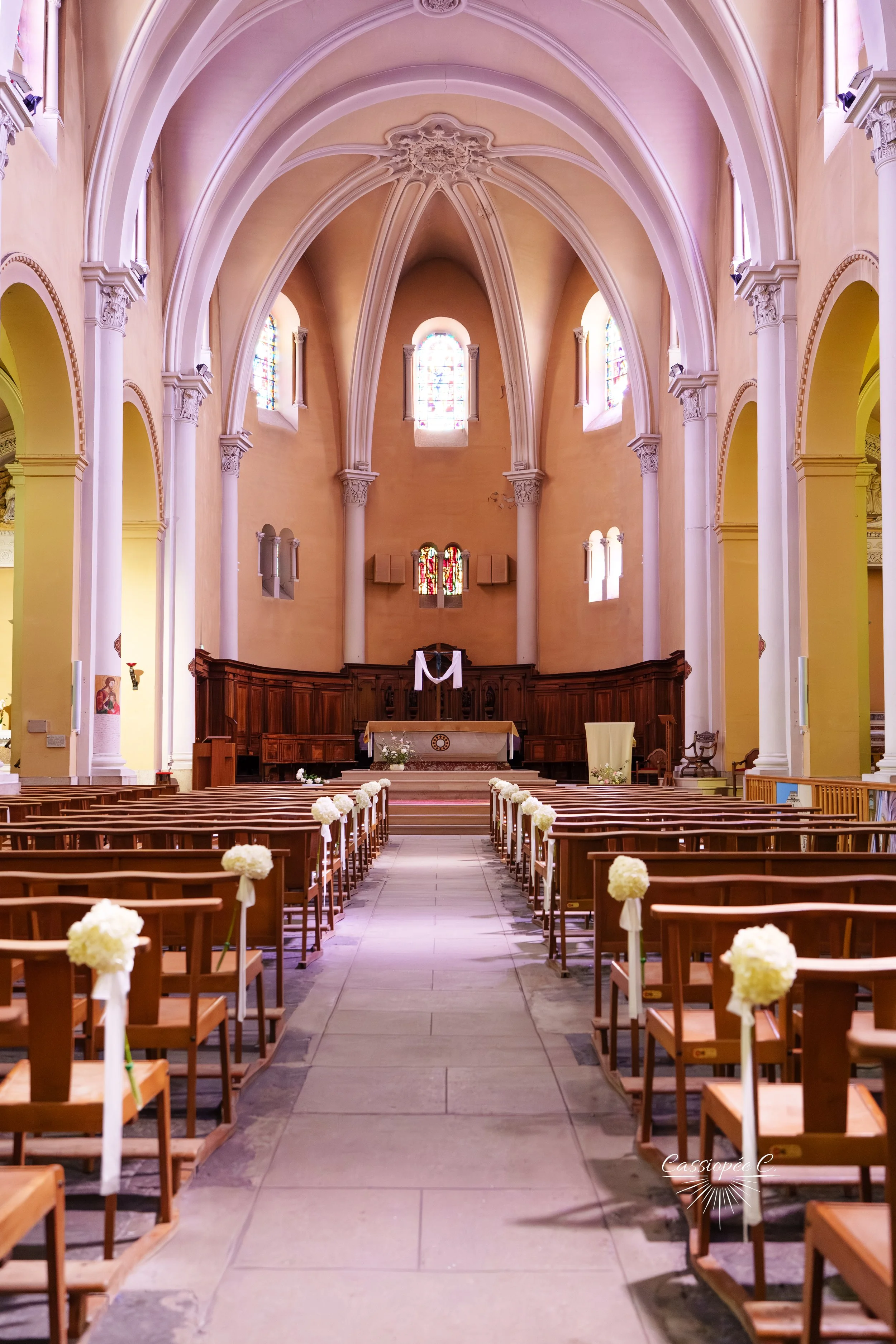 Intérieur d'une église aménagée pour un mariage, avec des chaises en bois décorées de bouquets de fleurs blanches le long d'une allée centrale menant à l'autel.