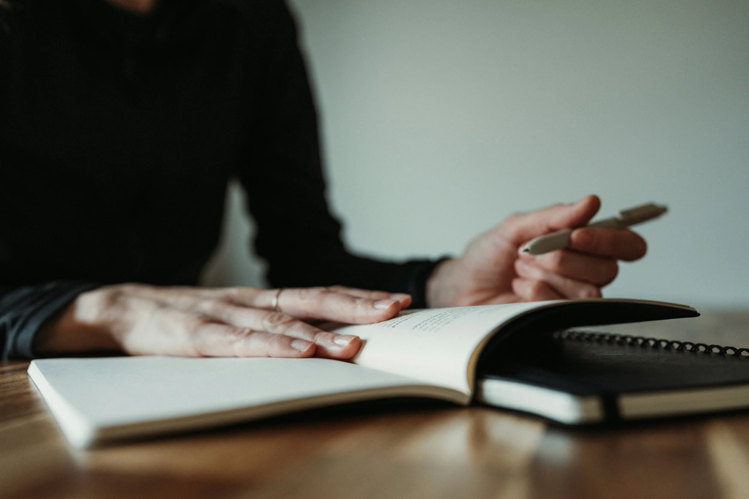 A person wearing a black long sleeve shirt is sitting at a wooden table, writing in a notebook with a pen, with an open book and additional notebooks nearby.