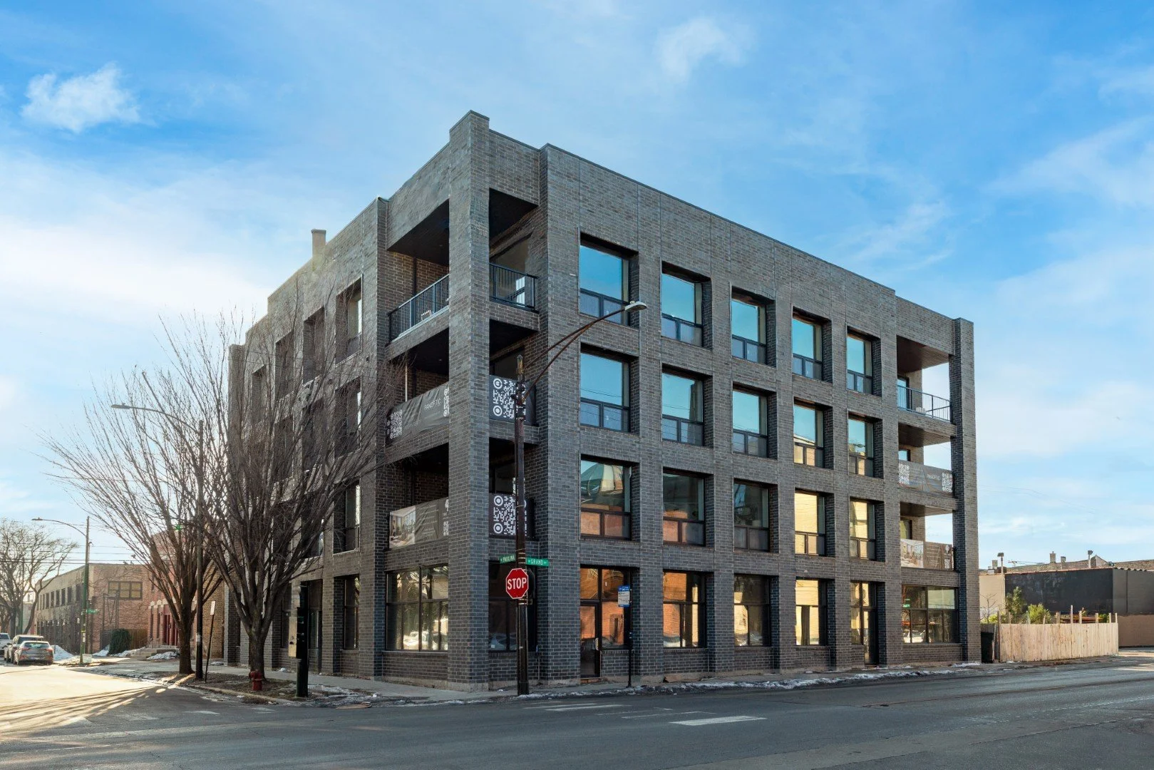 Modern 4-story apartment building with dark brick exterior, large windows, and small balconies at each level, situated on a city street with trees and cars.