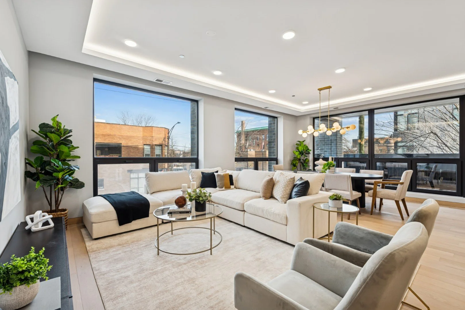 Modern living room with large windows, beige curtains, brown sofa with pillows, glass-top coffee tables, indoor plants, and a dining area with a chandelier.