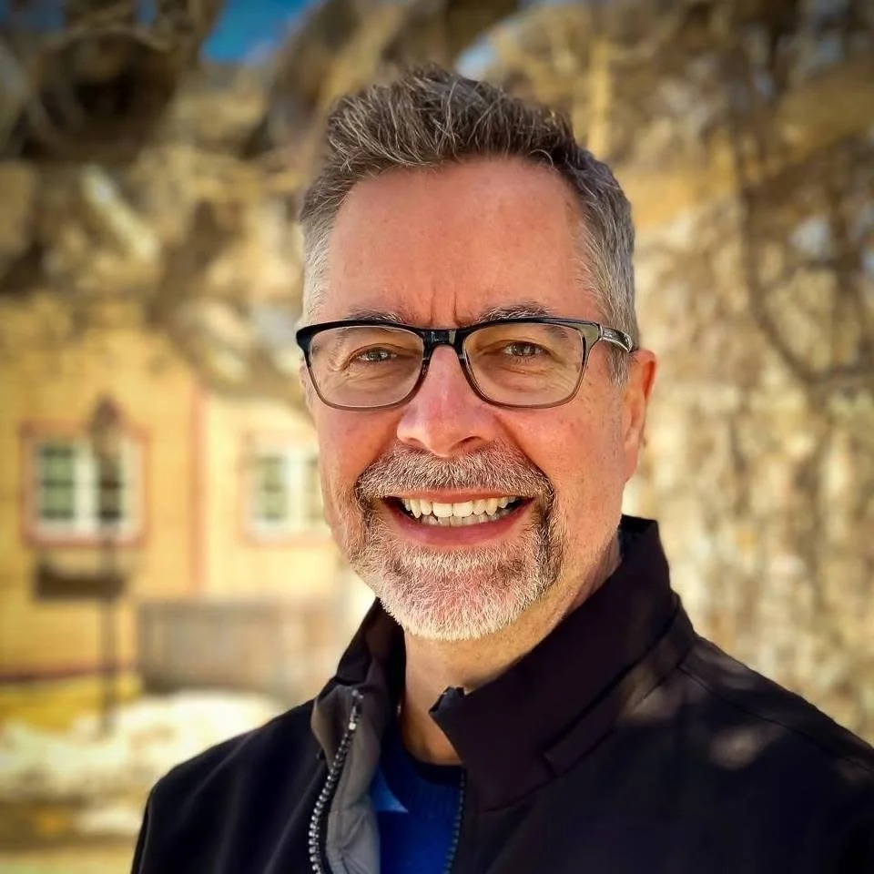 A middle-aged man with glasses, gray hair, and a goatee smiling in front of a tree with a yellow building in the background.