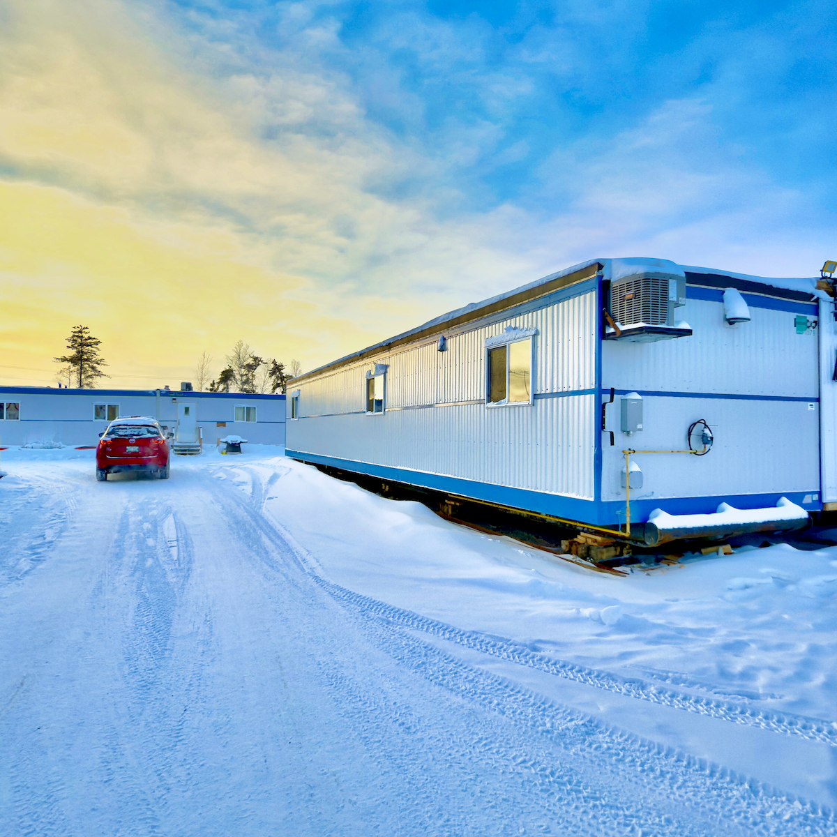 Snow-covered mobile home park with a red car, mobile trailers, and a bright sky.