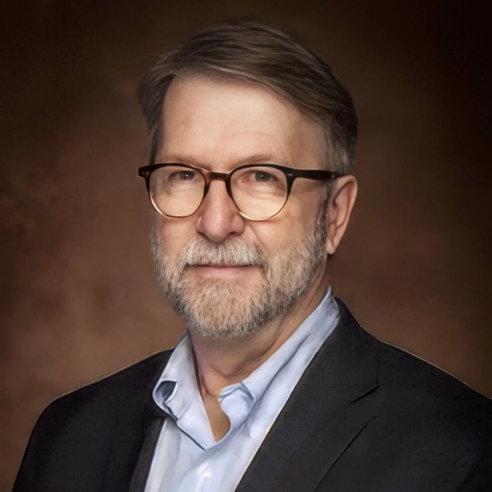 A middle-aged man with glasses, a beard, wearing a suit jacket and a light blue shirt, posing against a brown background.