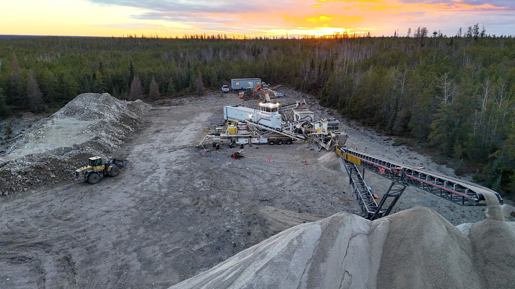 Mining site with a large machine processing gravel, a conveyor belt, a loader, and parked vehicles in a cleared area surrounded by forest, sunset in the background.