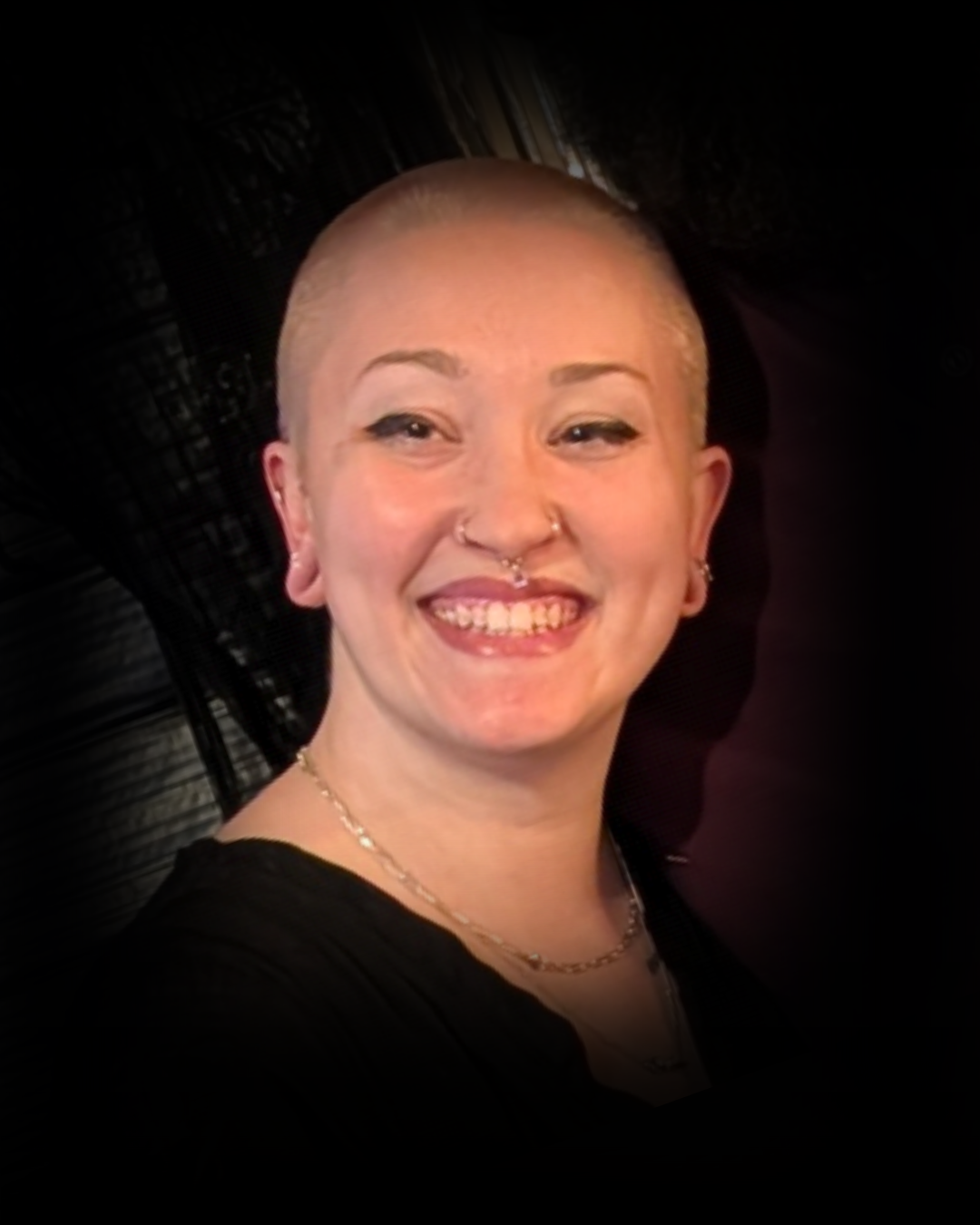 A woman with a shaved head smiling, wearing earrings, a nose ring, a necklace, and a black top.