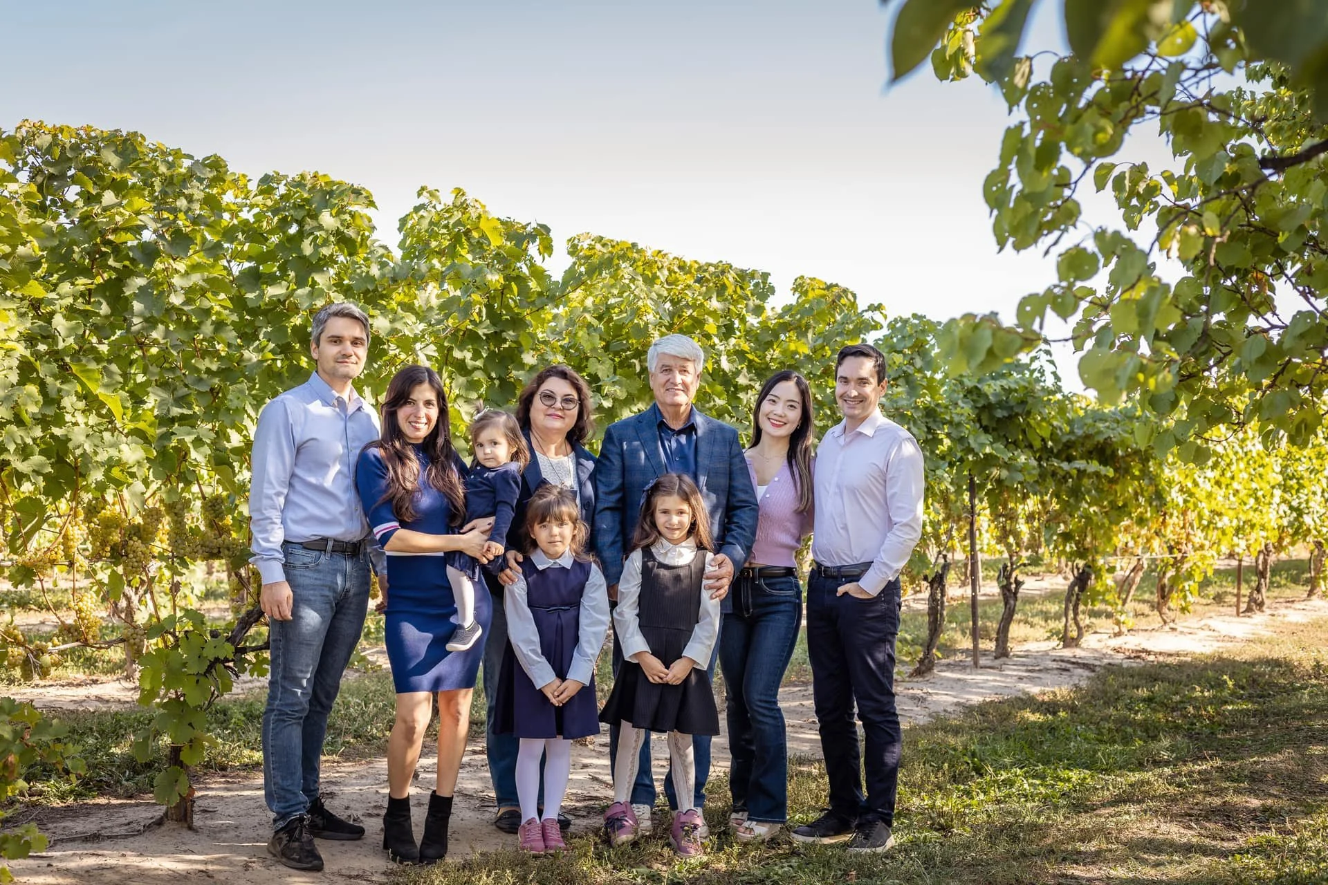 Family photo of nine people standing in a vineyard with grapevines in the background on a sunny day.