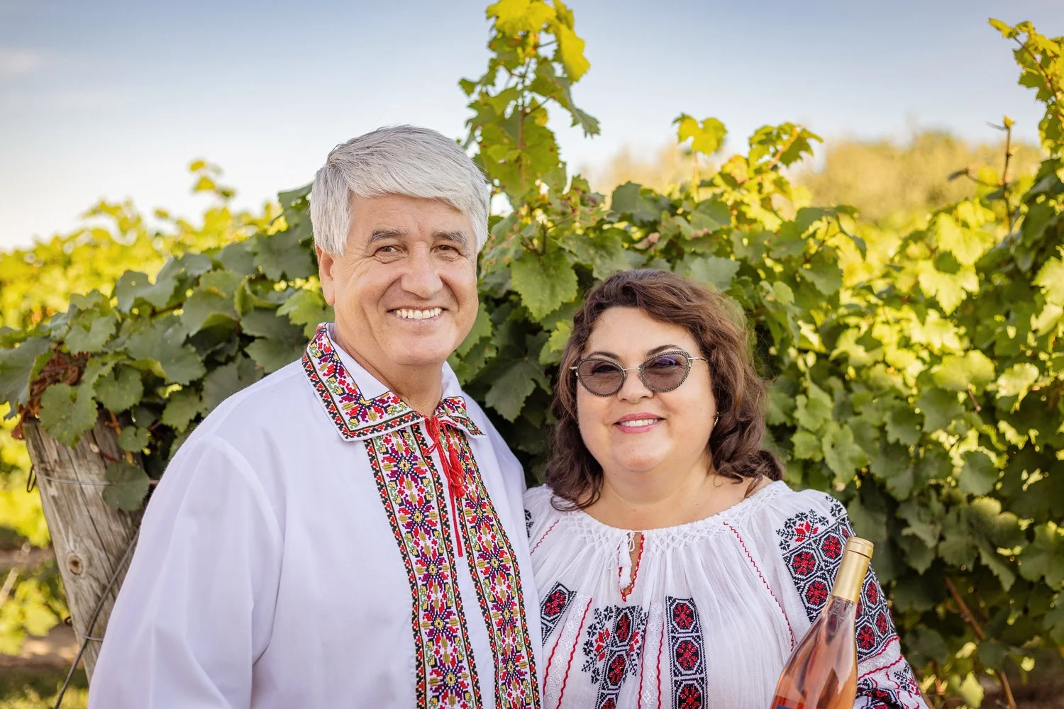 A smiling man and woman in traditional embroidered Moldovan clothing standing in a vineyard with green grapevines in the background. The woman is wearing sunglasses and holding a bottle of rosé wine.