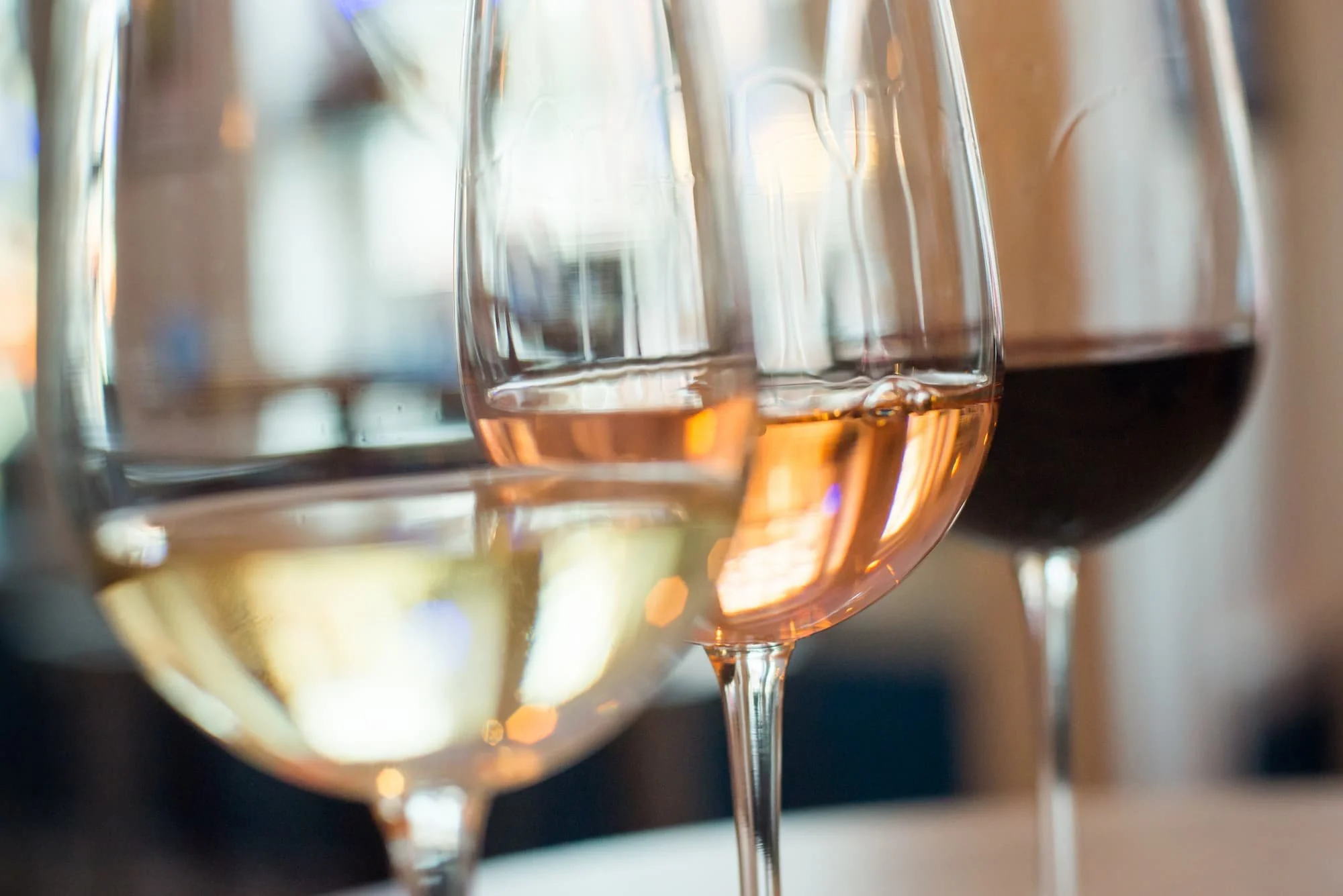 Close-up of three wine glasses, containing white, rosé, and red wine, on a table with blurred background.