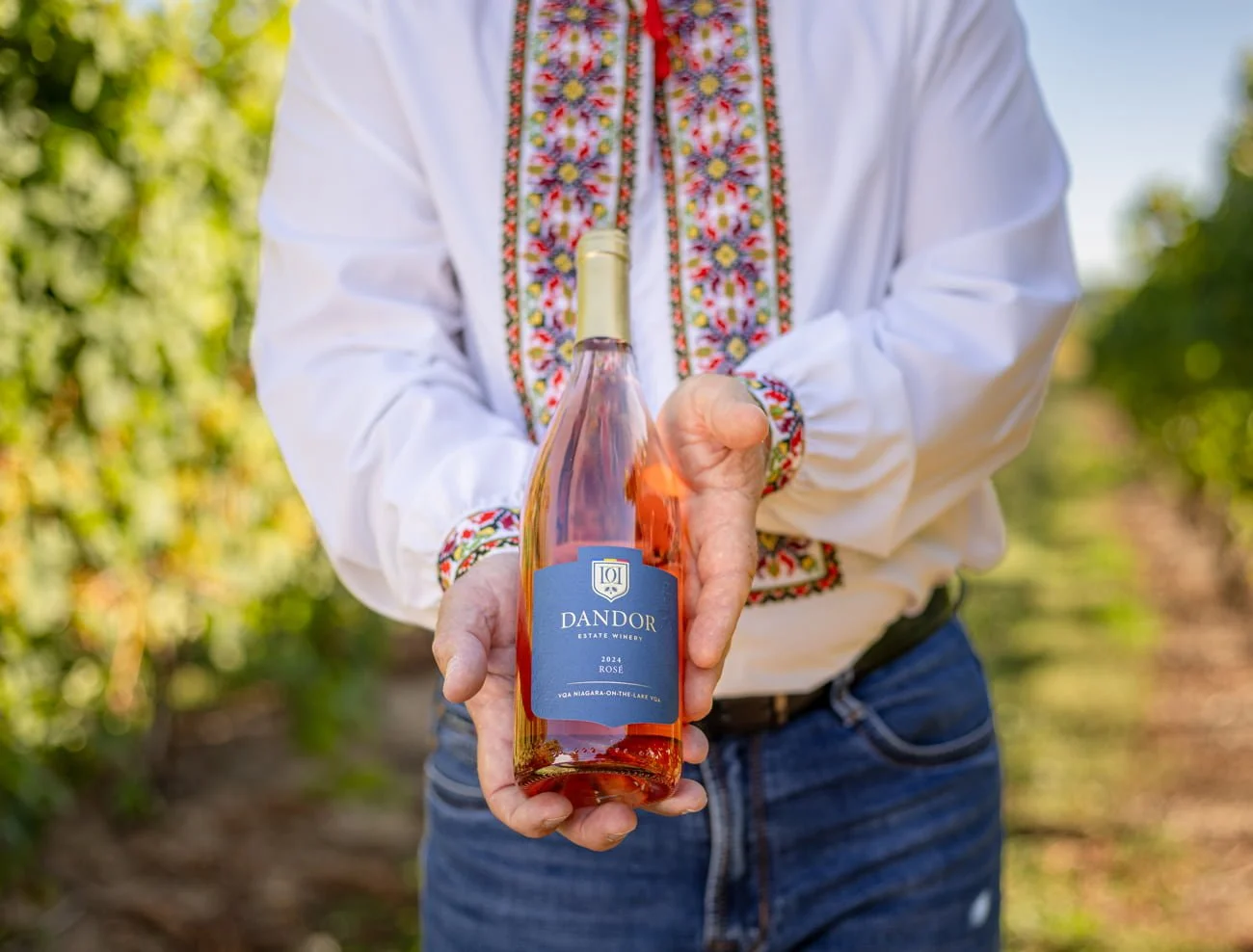 Traditional embroidered shirt holding a bottle of rosé wine in a vineyard.