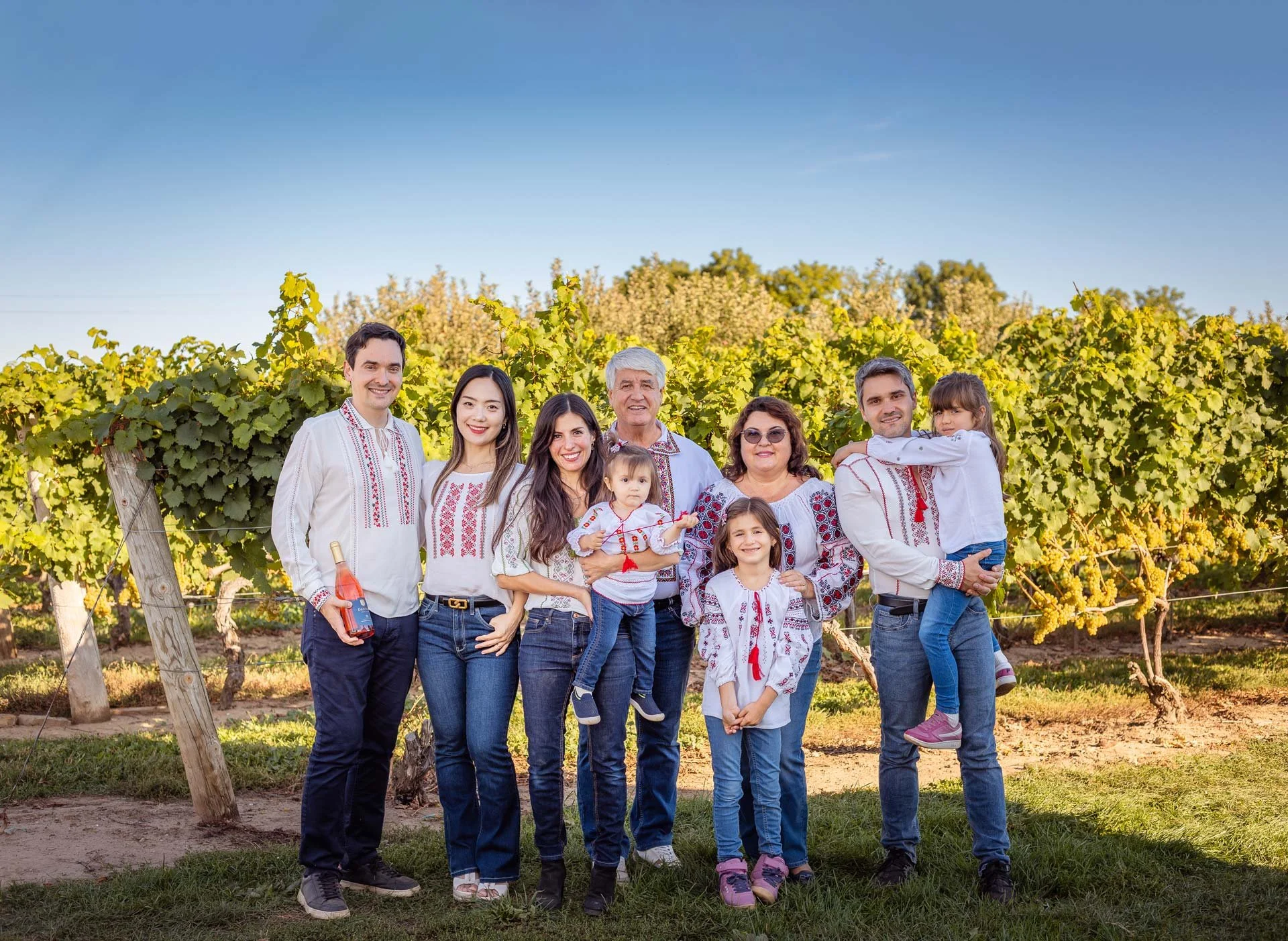 Family of ten posing in a vineyard during daytime, all wearing traditional embroidered clothing, with green vines and a blue sky in the background.