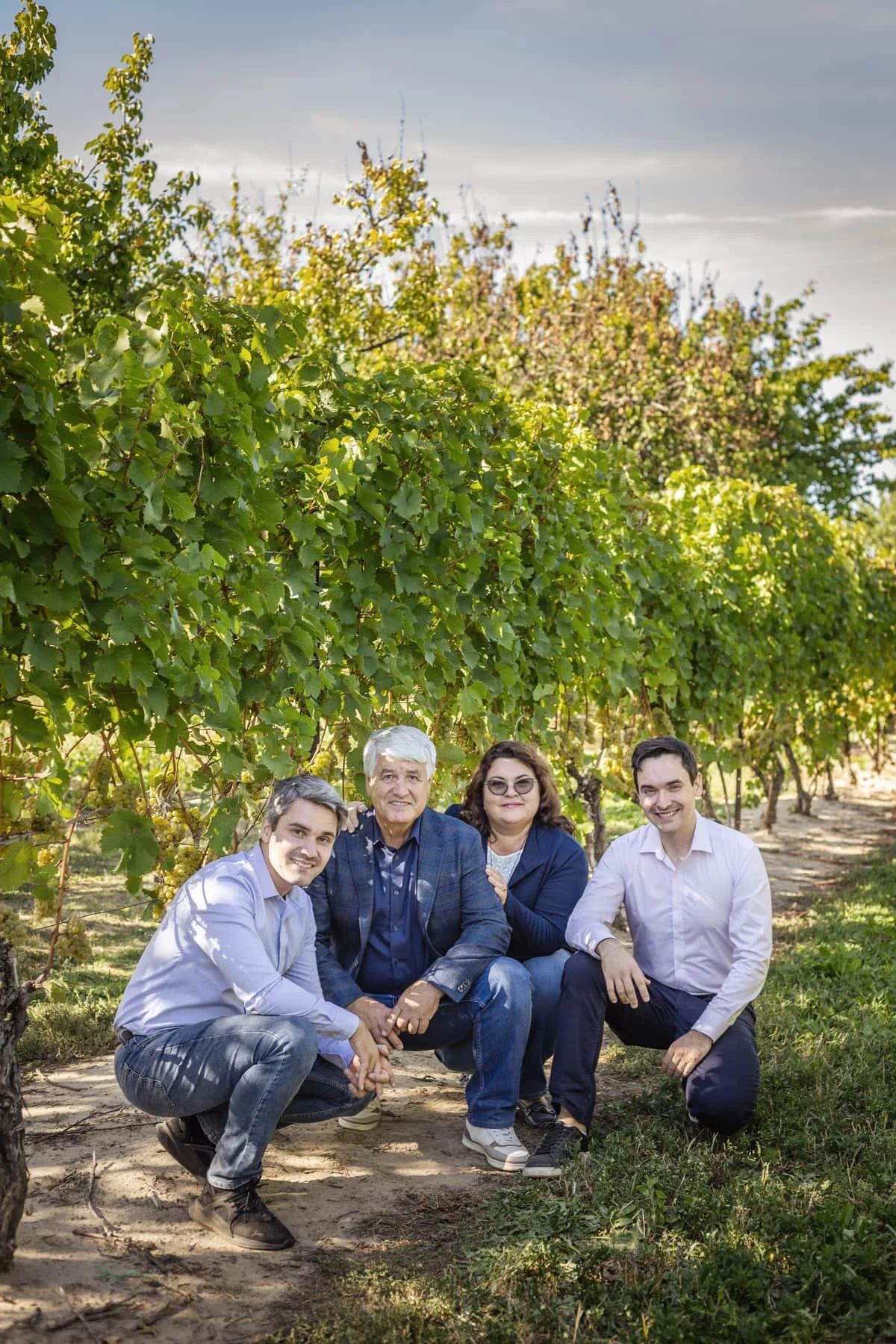 Four people, two men and two women, are crouching and kneeling in a vineyard with green grapevines behind them.