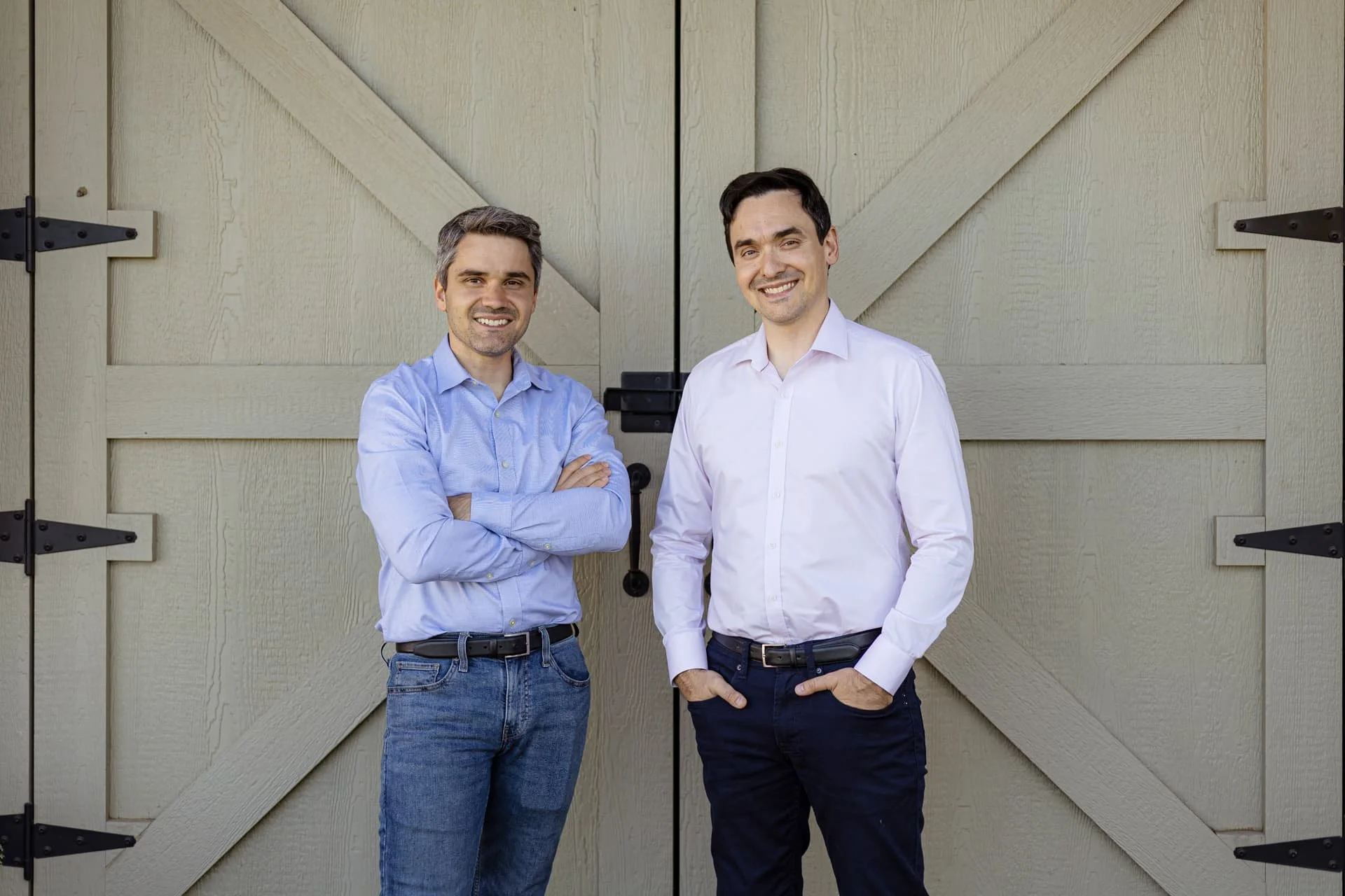 Two men standing in front of a wooden barn door, smiling. One man has gray hair and is wearing a light blue shirt with jeans; the other has dark hair and is wearing a white shirt with dark pants.