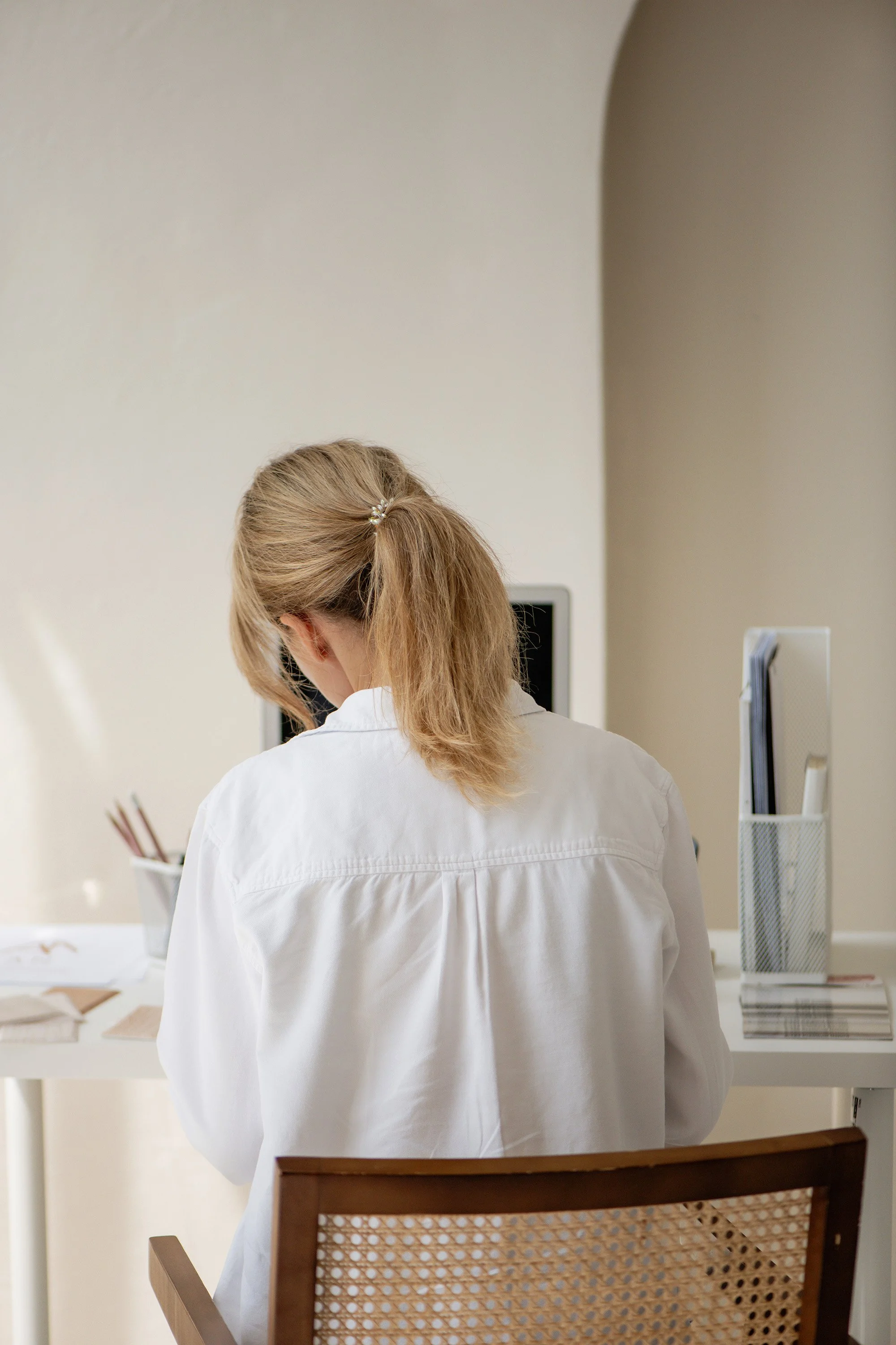 Woman with blonde hair tied in a ponytail sitting at a white desk, facing away, in a minimal office space.