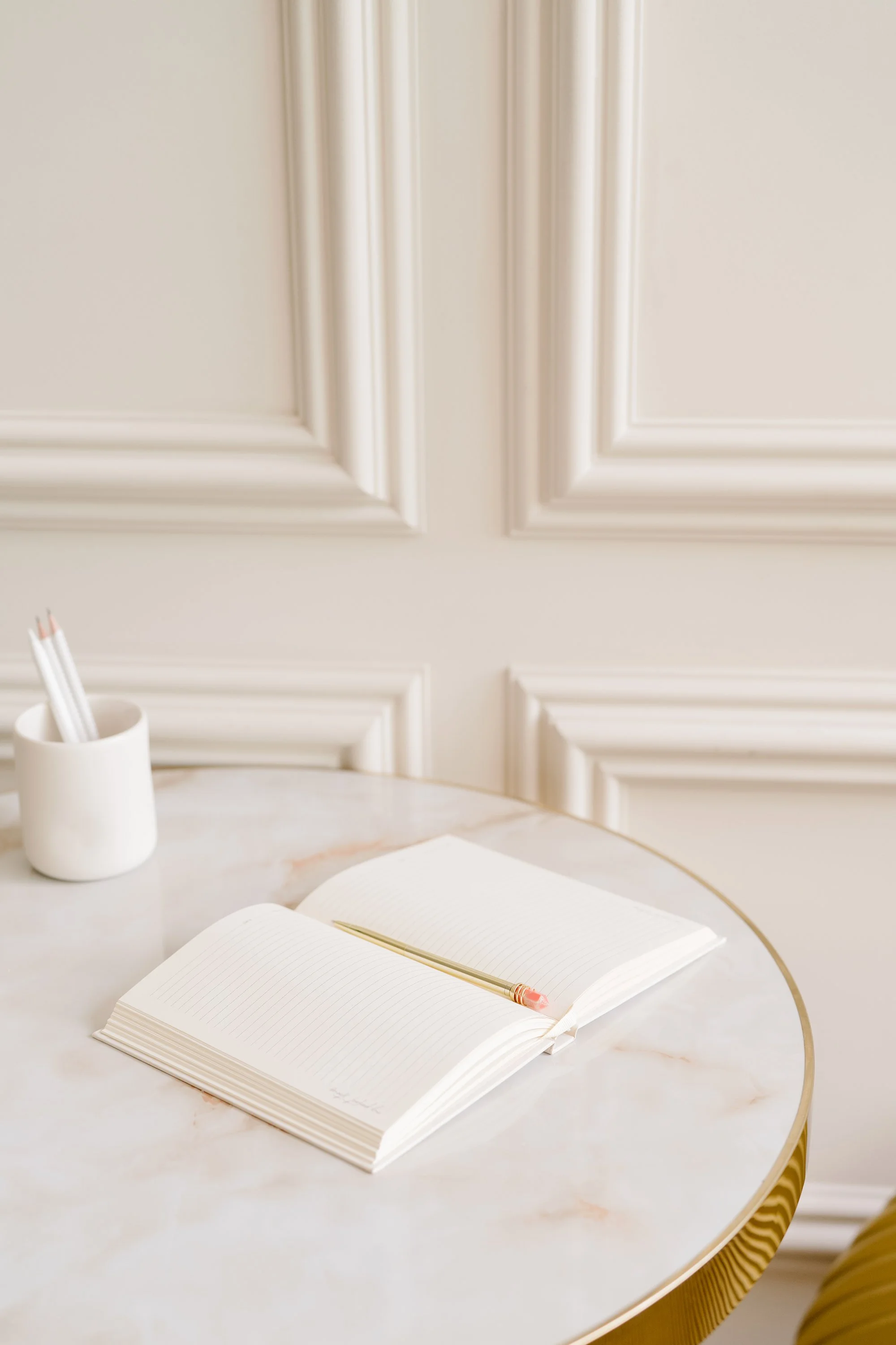 Open white notebook with a gold pen on a marble table next to a white cup holding white pencils, with a cream-colored decorative wall in the background.