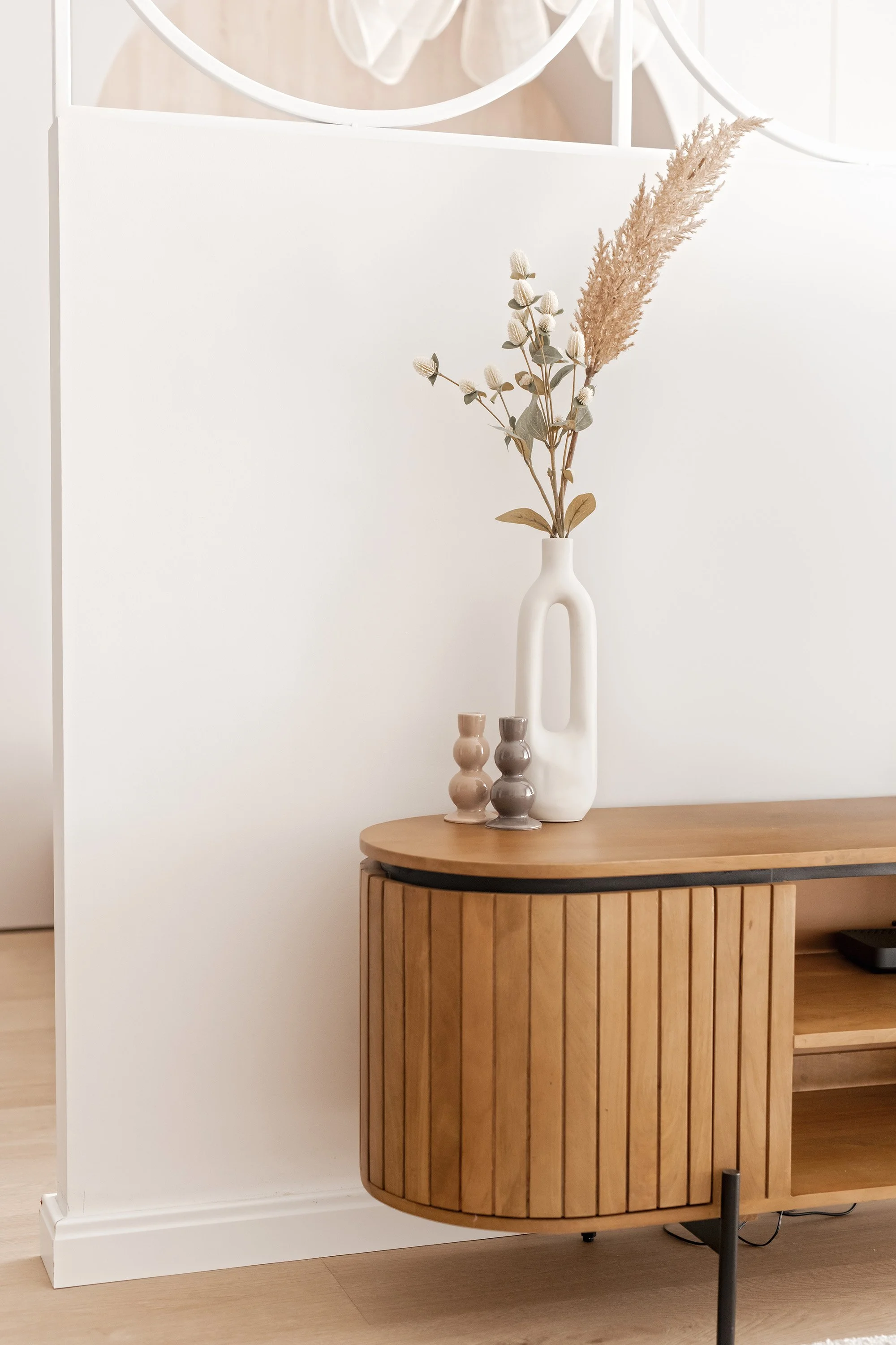 A white ceramic vase with an elongated handle contains dried pampas grass and eucalyptus stems. Two small beige and gray ceramic candle holders are beside the vase. The arrangement sits on a wooden sideboard with a curved front and vertical slats, against a white wall in a modern, minimalist interior.