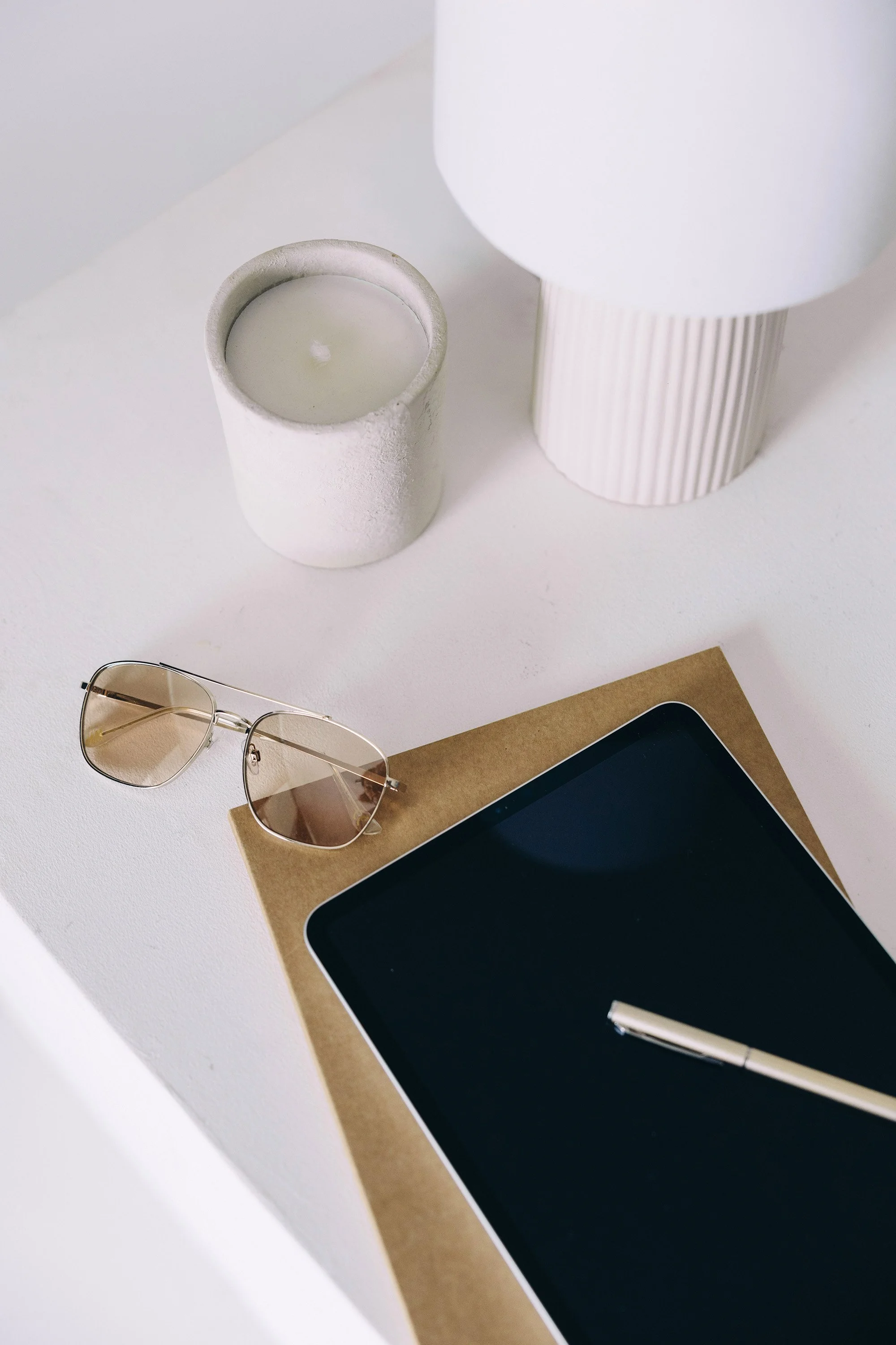 A white desk with a pair of sunglasses, a tablet with a black screen and white border, a gold pen on top, a brown paper folder, a white candles in a container, and a textured white table lamp.