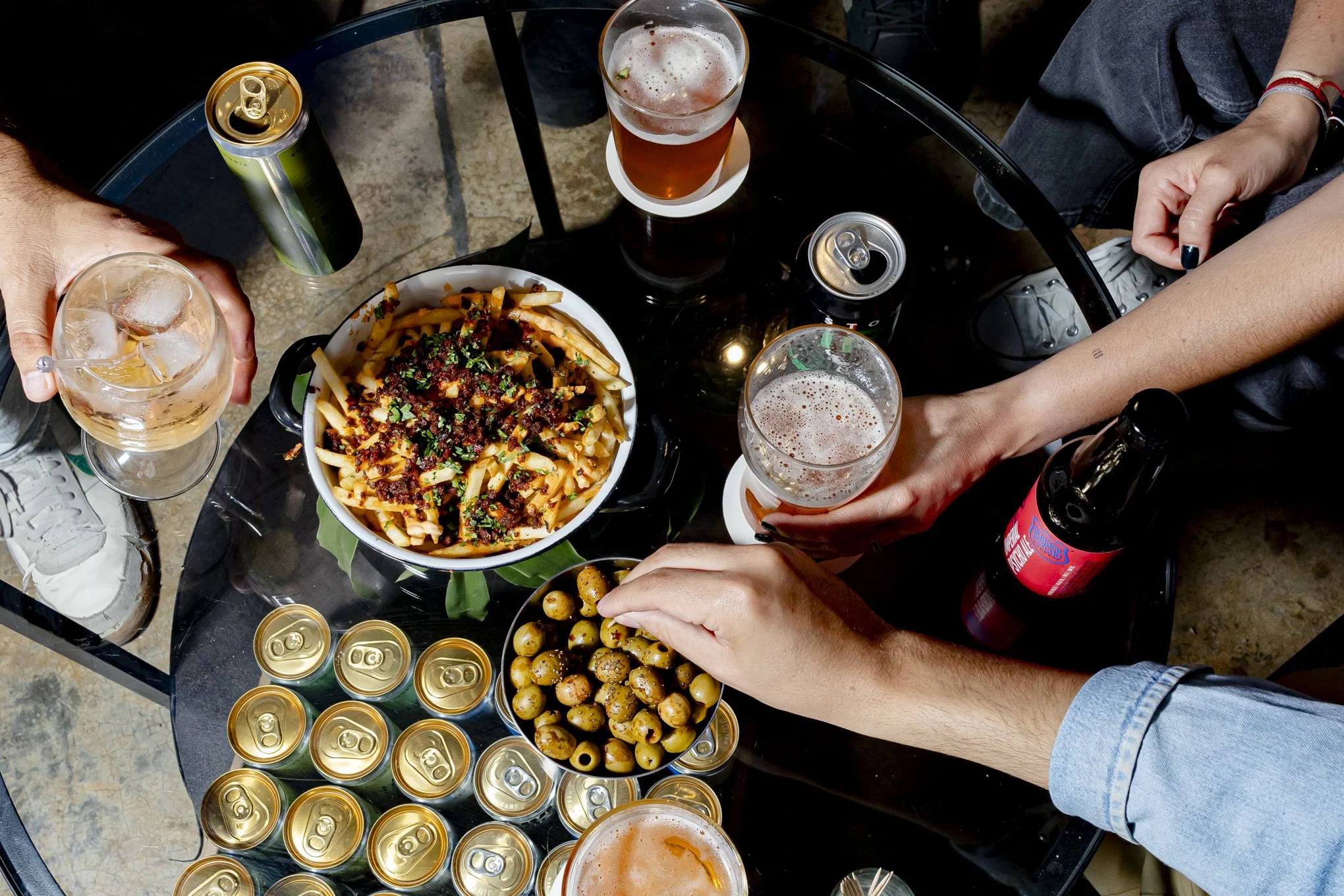 Un grupo de personas compartiendo una comida y bebidas en una mesa redonda de cristal, con un plato de pasta, varias latas de bebidas, y varias copas con cerveza en un ambiente informal.