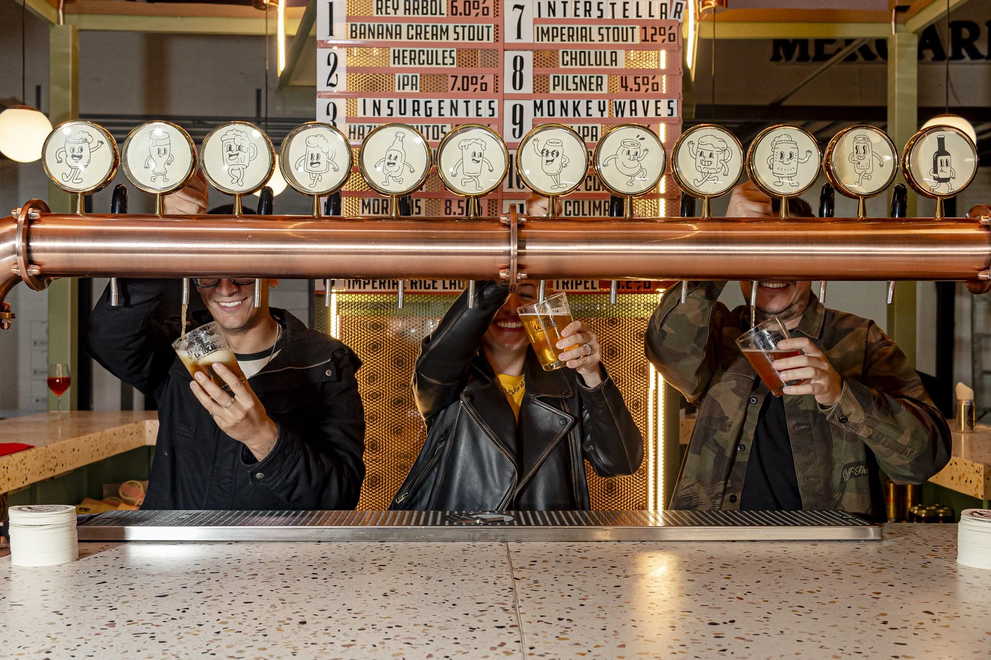Tres personas en un bar, levantando copas de cerveza y sonriendo, con un cilindro de cobre con etiquetas de dibujos animados y un cartel de cervezas en el fondo.