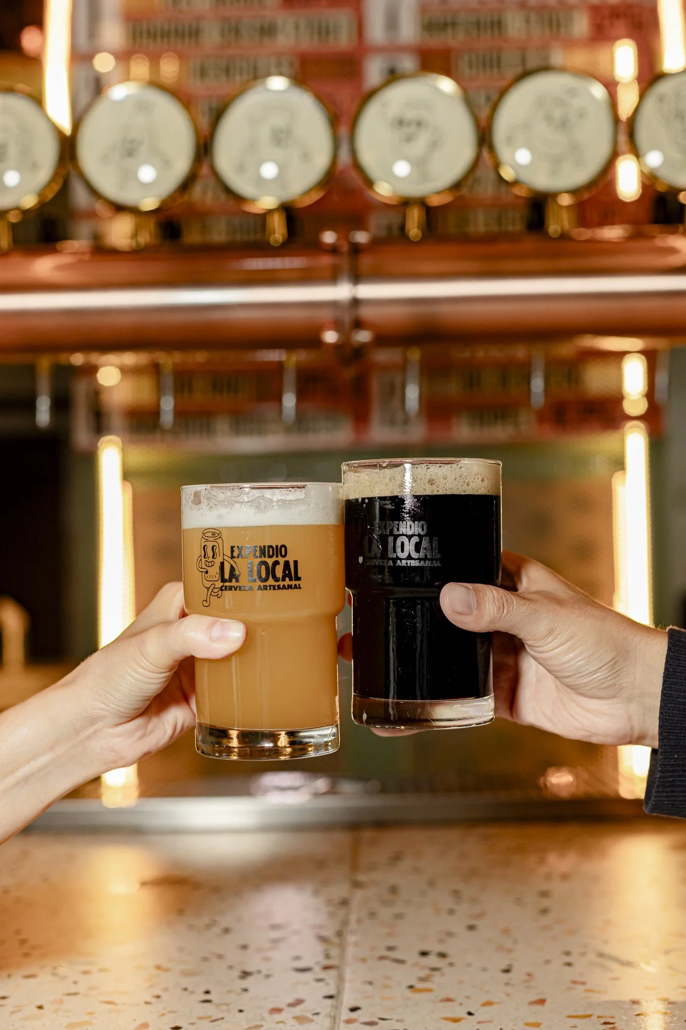 Dos manos sosteniendo vasos de cerveza artesanal, una con cerveza clara y otra con cerveza negra en un bar.