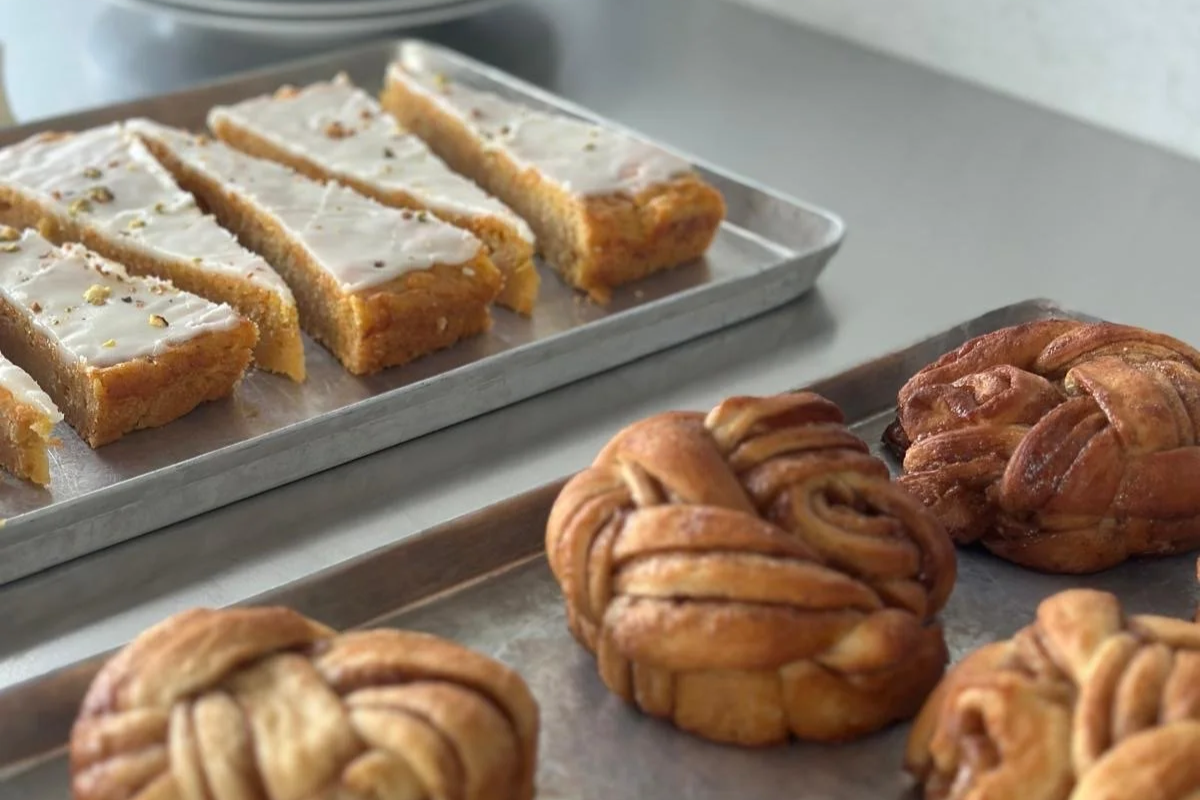 Vegane Zitronenriegel mit weißer Glasur und essbaren Blüten, sowie Knotensschnecken mit Zimt und Zucker auf Backblechen.