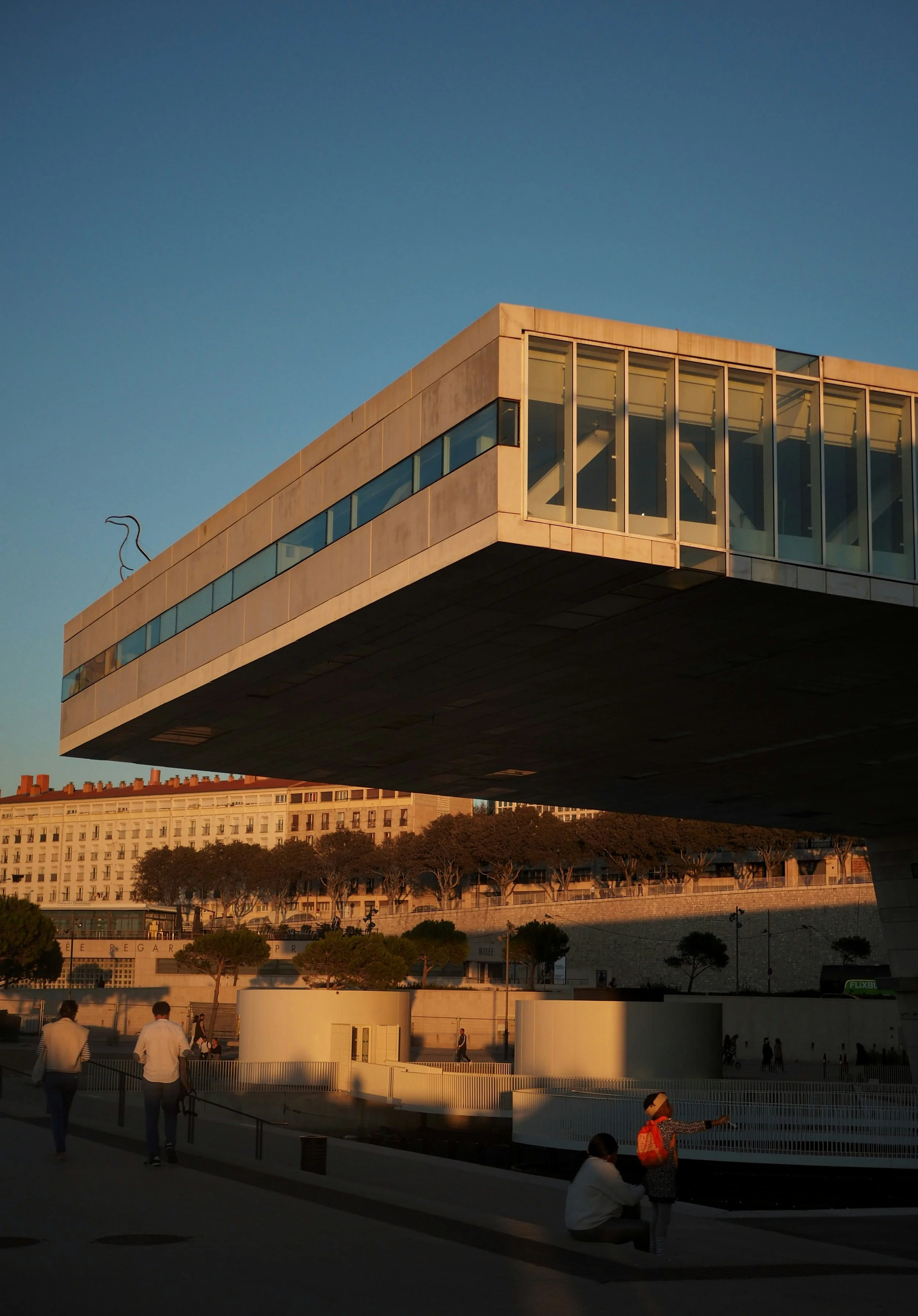 Vue d'un bâtiment moderne avec une structure suspendue, ainsi que des personnes dans une zone urbaine au coucher du soleil.