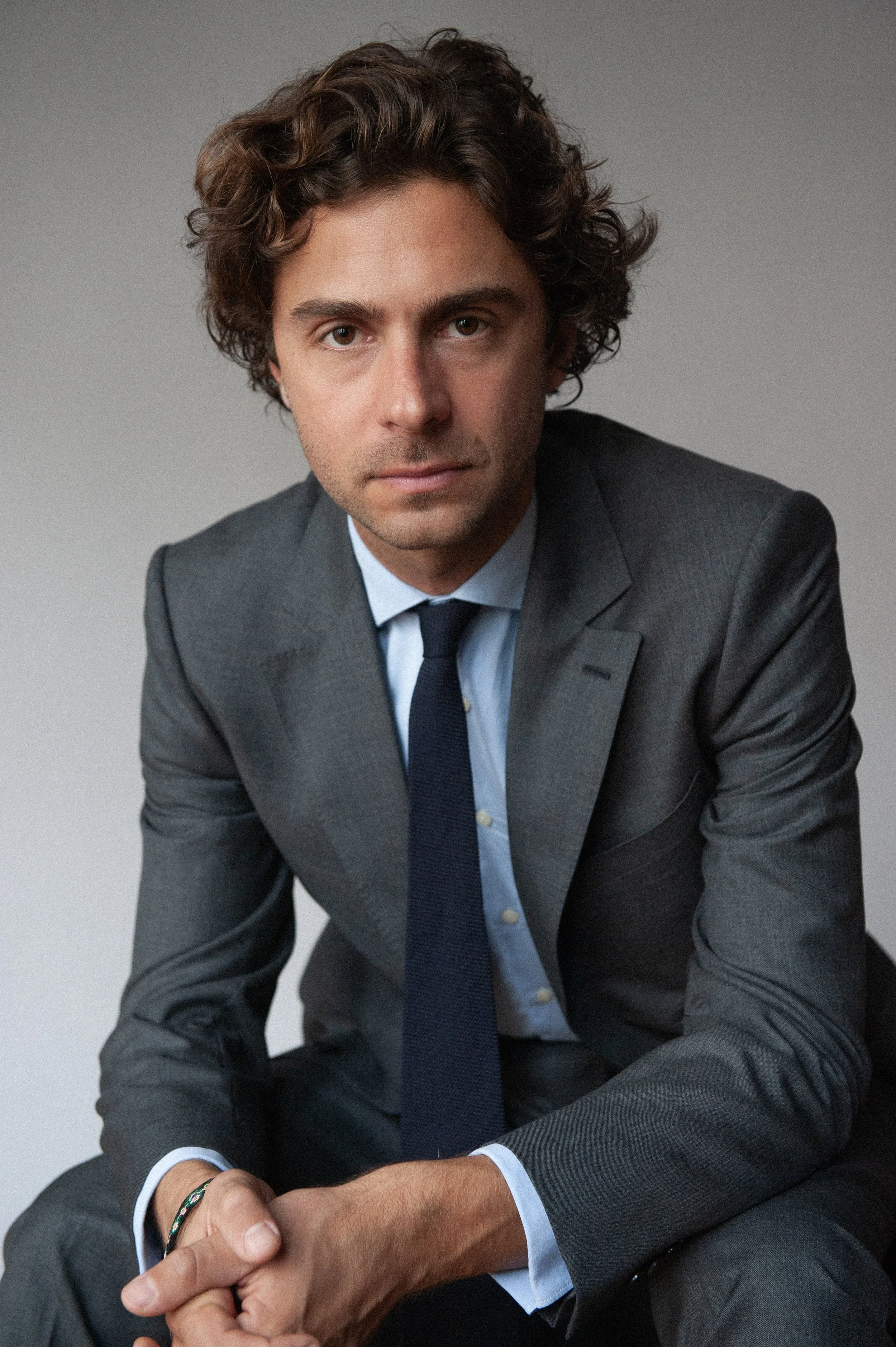 Jeune homme en costume gris, veste boutonnée, chemise bleue et cravate, assis contre un fond gris clair.