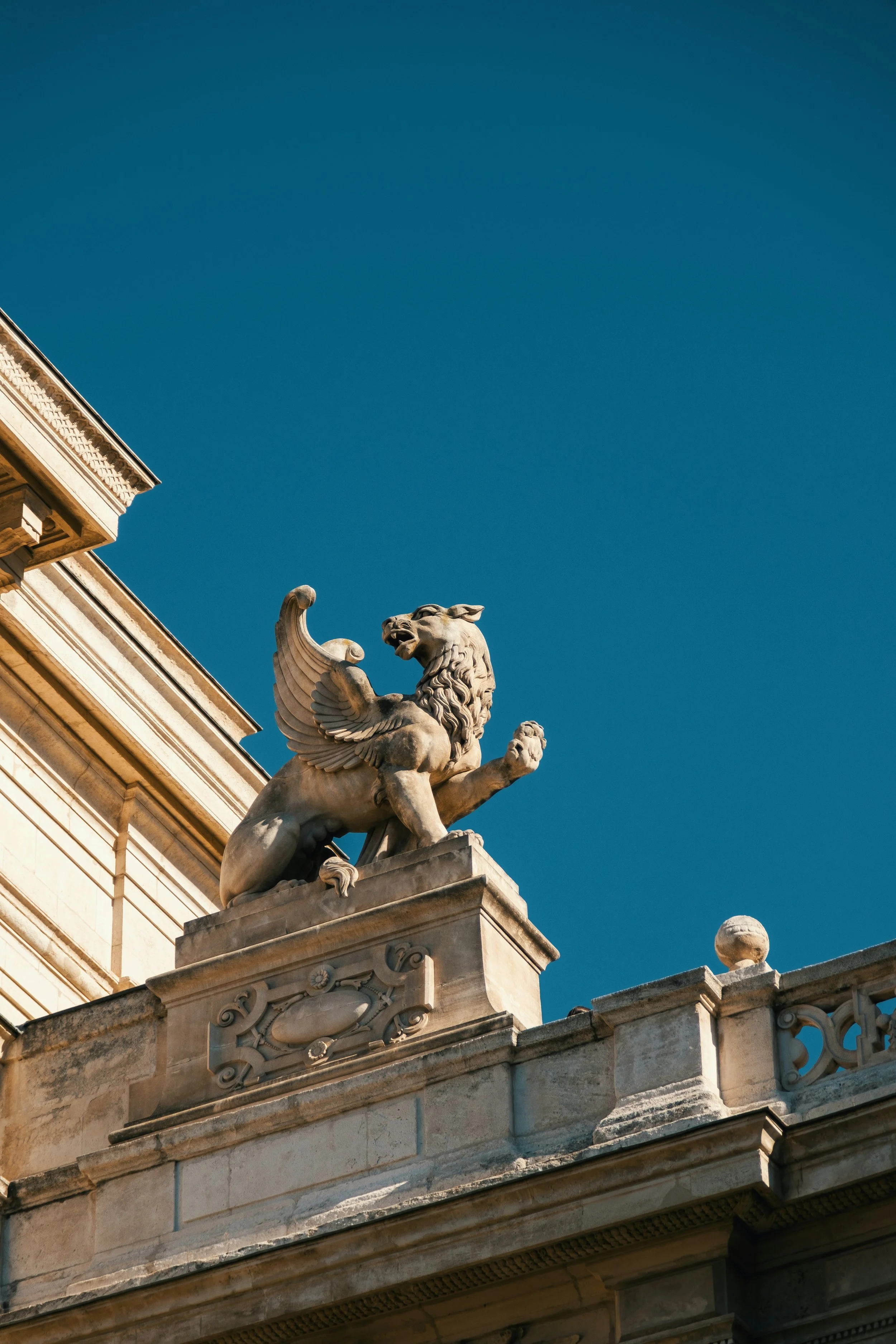 Une sculpture en pierre d'un lion avec des ailes, située en haut d'une balustrade d'un bâtiment historique, contre un ciel bleu clair.