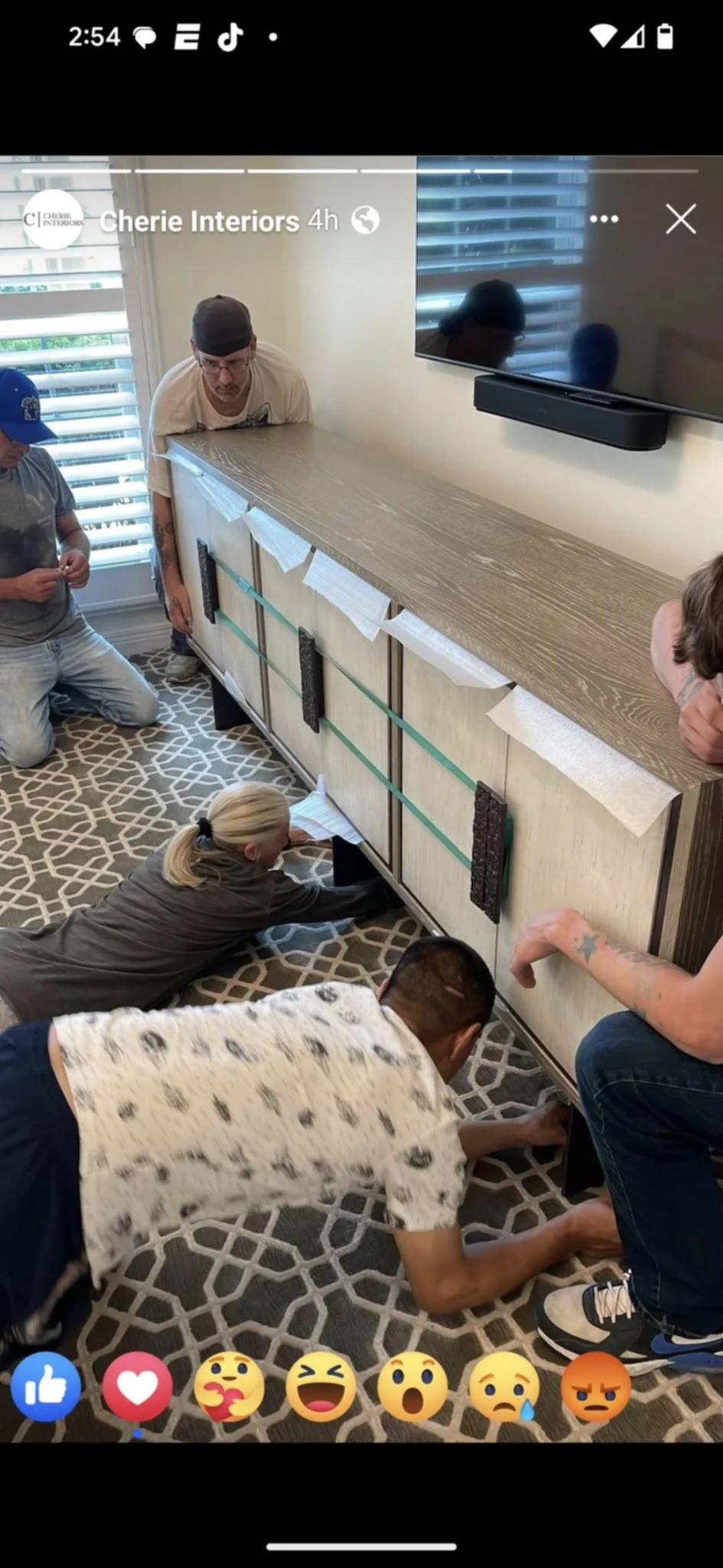 Group of people working together on furniture assembly in a living room, with one woman on the floor, some leaning on the cabinet, and others kneeling or sitting around, assembling or inspecting a large wooden cabinet against the wall. There is a tel