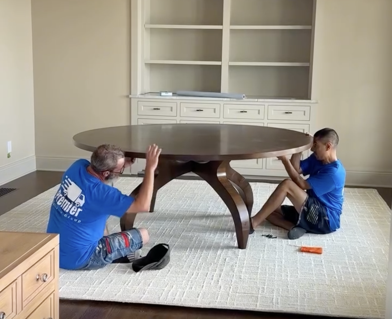 Two men dressed in blue, sitting on the floor under a large round wooden dining table, with one raising his hand and the other holding the table's edge, possibly assembling or fixing the table inside a room with a built-in white cabinet and shelves i