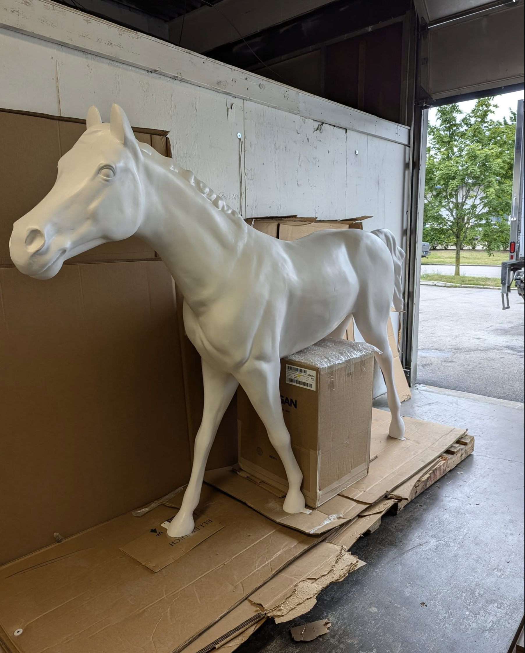 White horse sculpture in a warehouse surrounded by cardboard boxes and wood pallets, with a partially open garage door and trees outside.